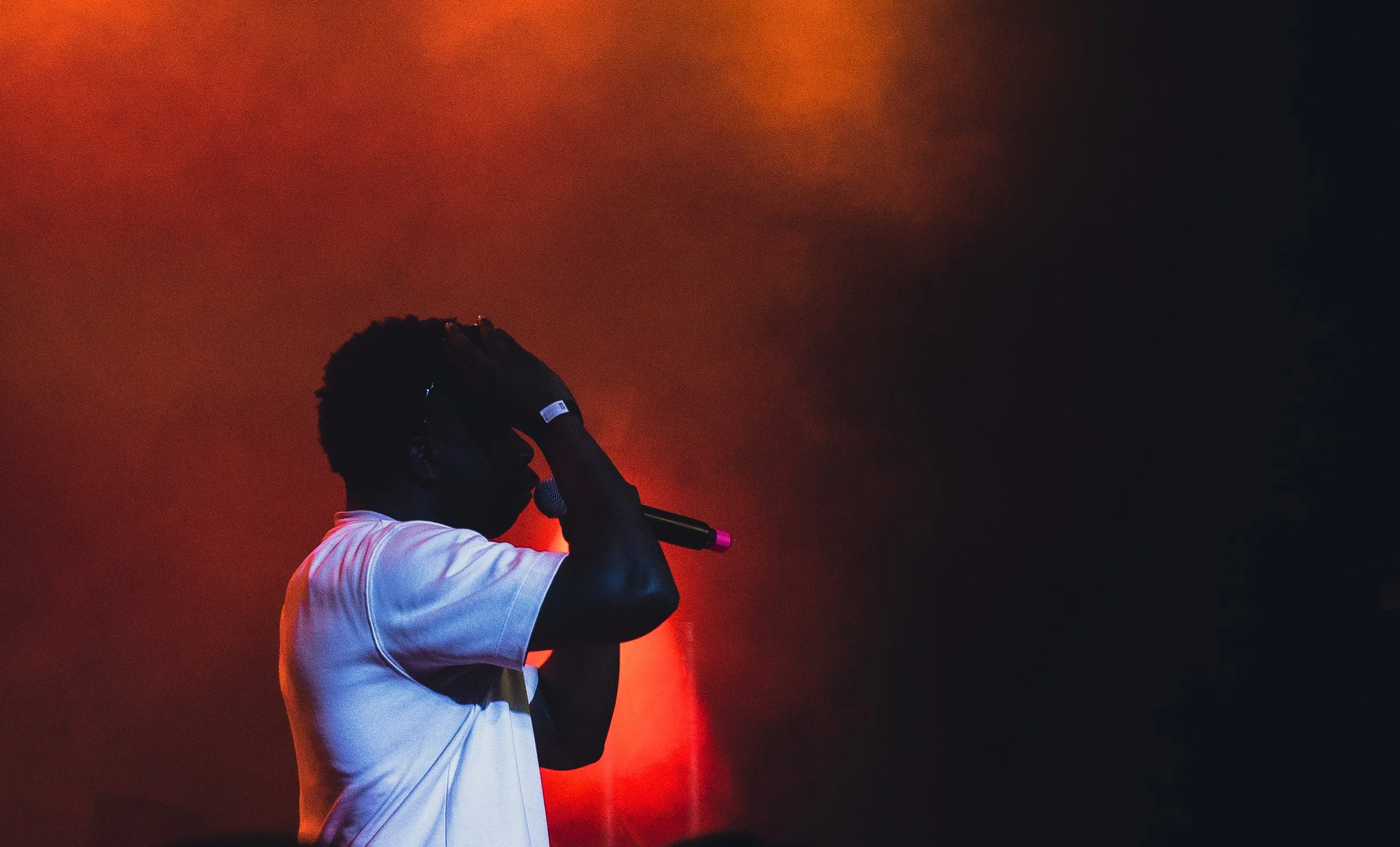 A person singing into a microphone on stage with dark background and red lighting.