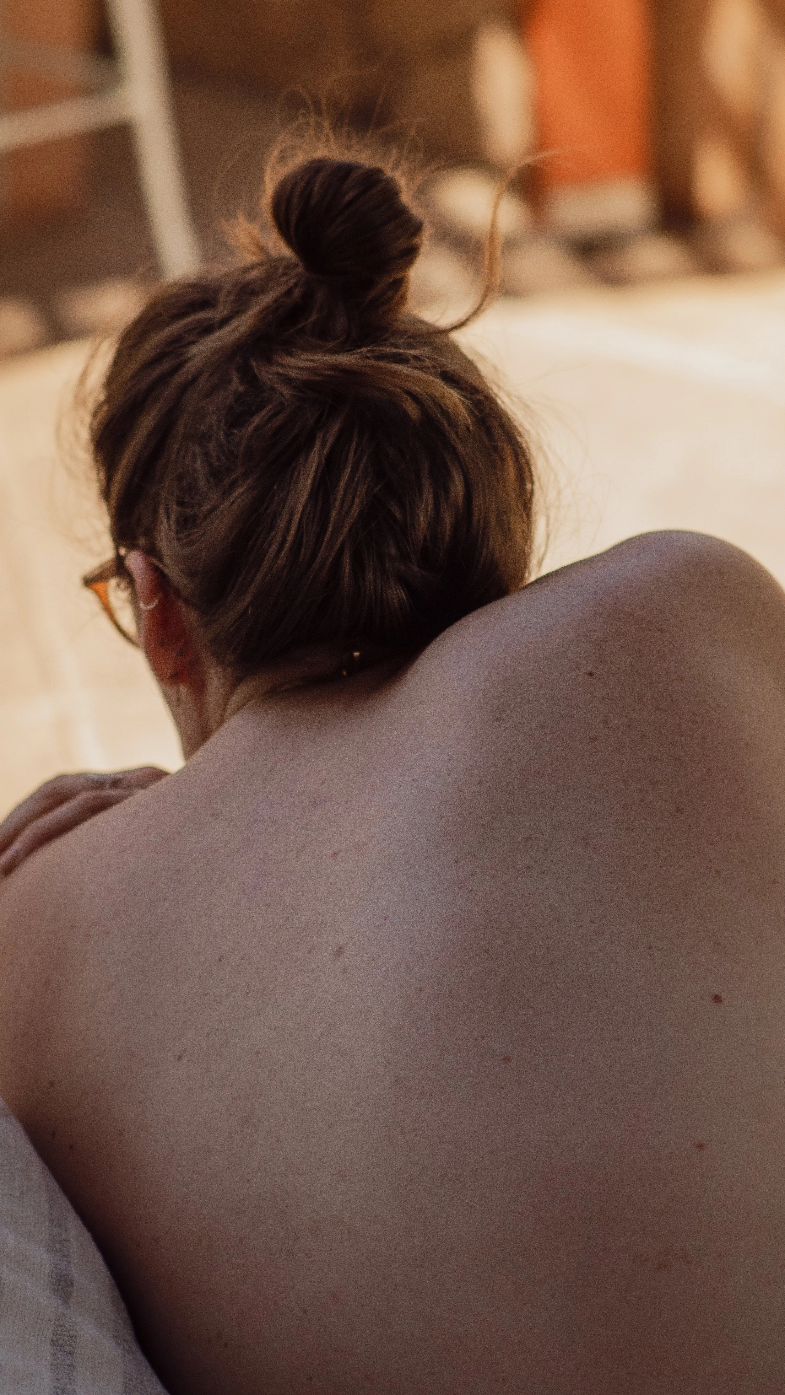 A person with red hair styled in a messy bun, resting their face on their arm, viewed from behind, showing their bare shoulder and back.