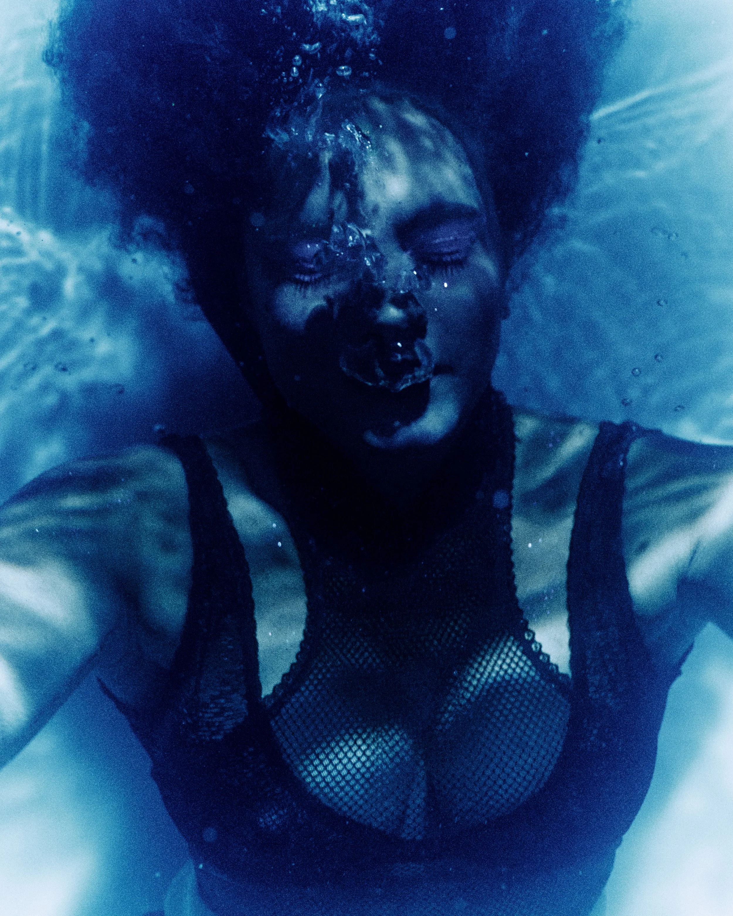 A woman submerged underwater with her eyes closed, wearing a black fishnet top, and her hair floating around her head.