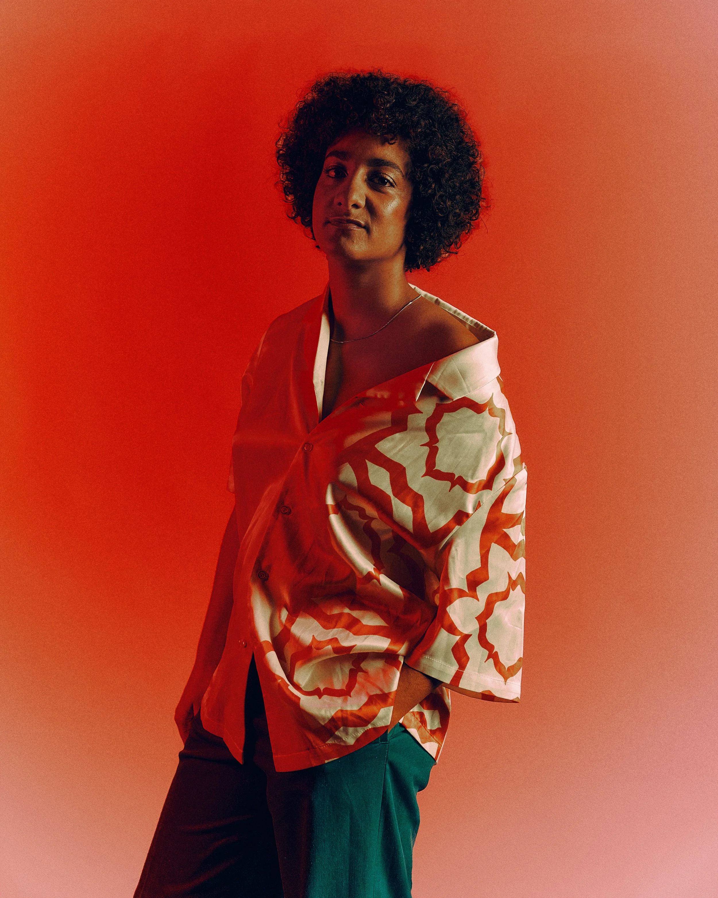 A woman with dark curly hair wearing a patterned button-up shirt with orange and white designs, standing against a red and orange gradient background.