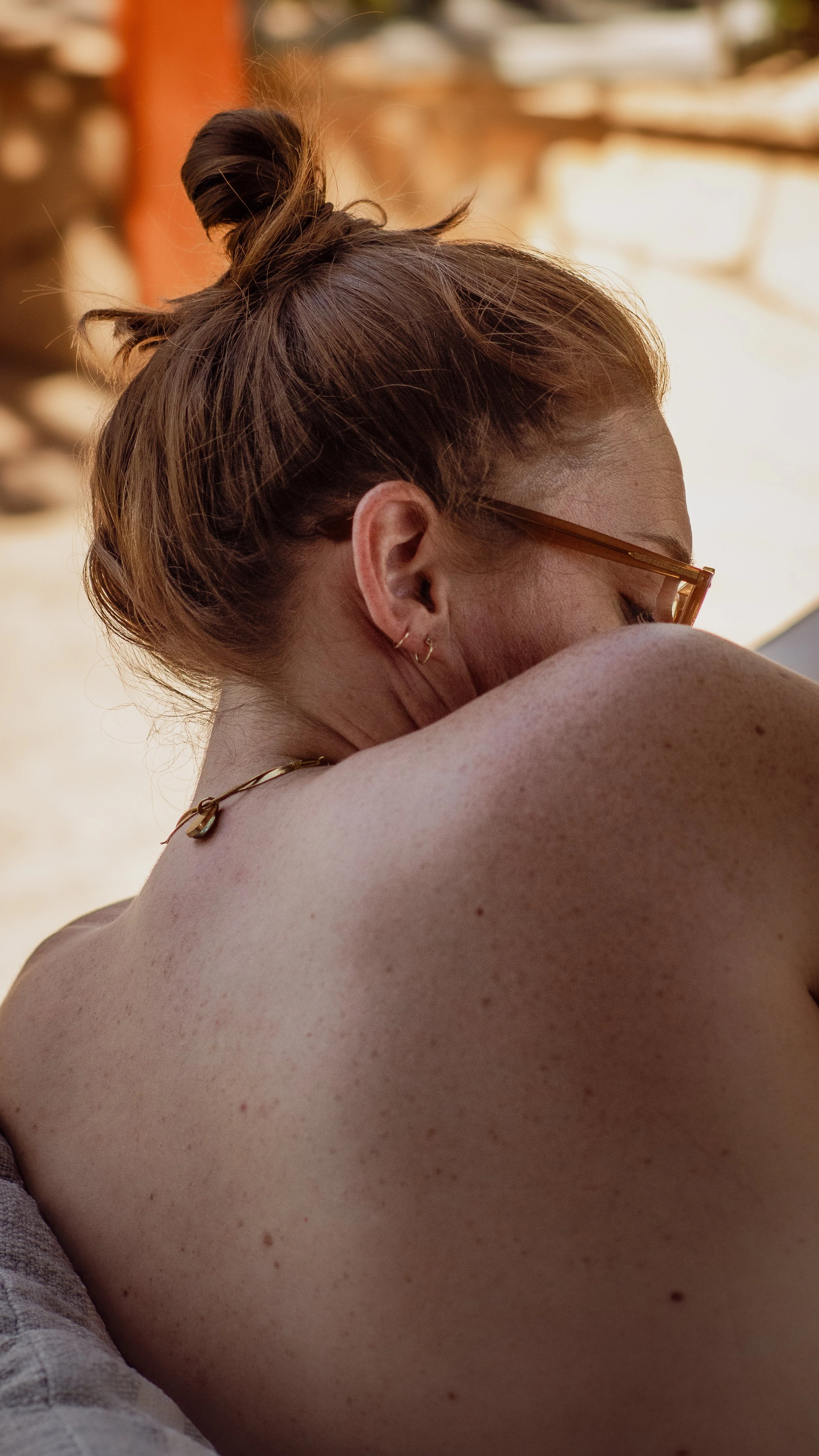A woman with red hair in a top bun, wearing glasses and multiple earrings, is resting her head on her shoulder in a relaxed outdoor setting.