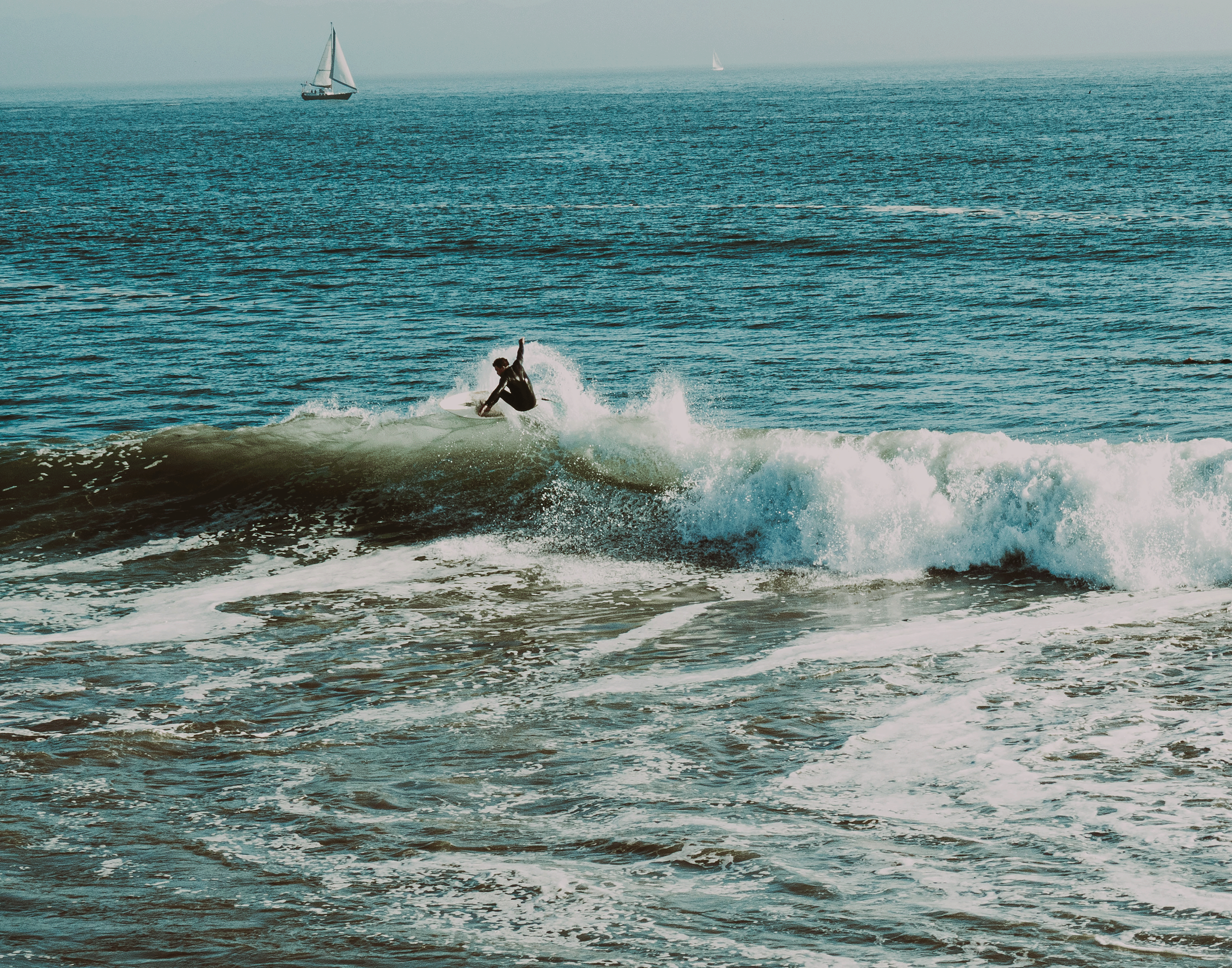 A person surfing on a wave in the ocean with sailboats in the distance.