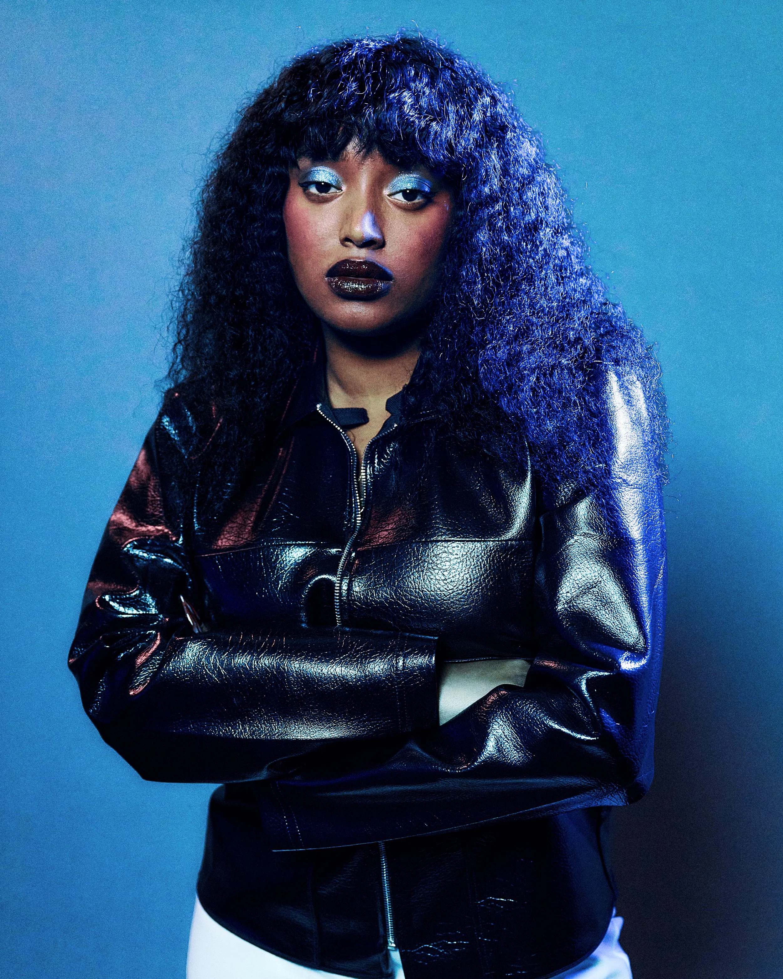 A woman with curly hair wearing a black shiny leather jacket with her arms crossed against a blue background.