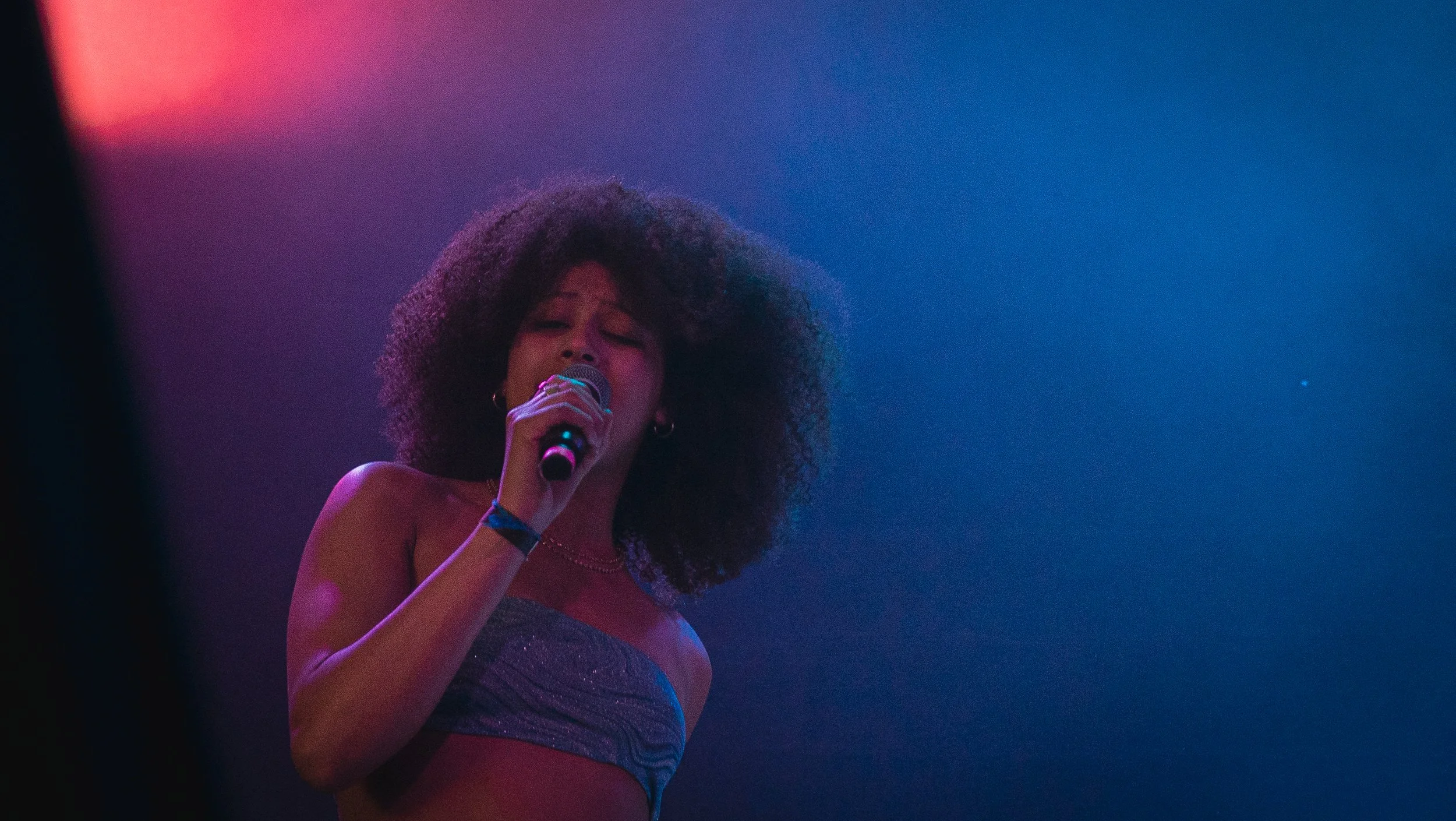 A woman with curly hair singing into a microphone with colorful stage lighting.