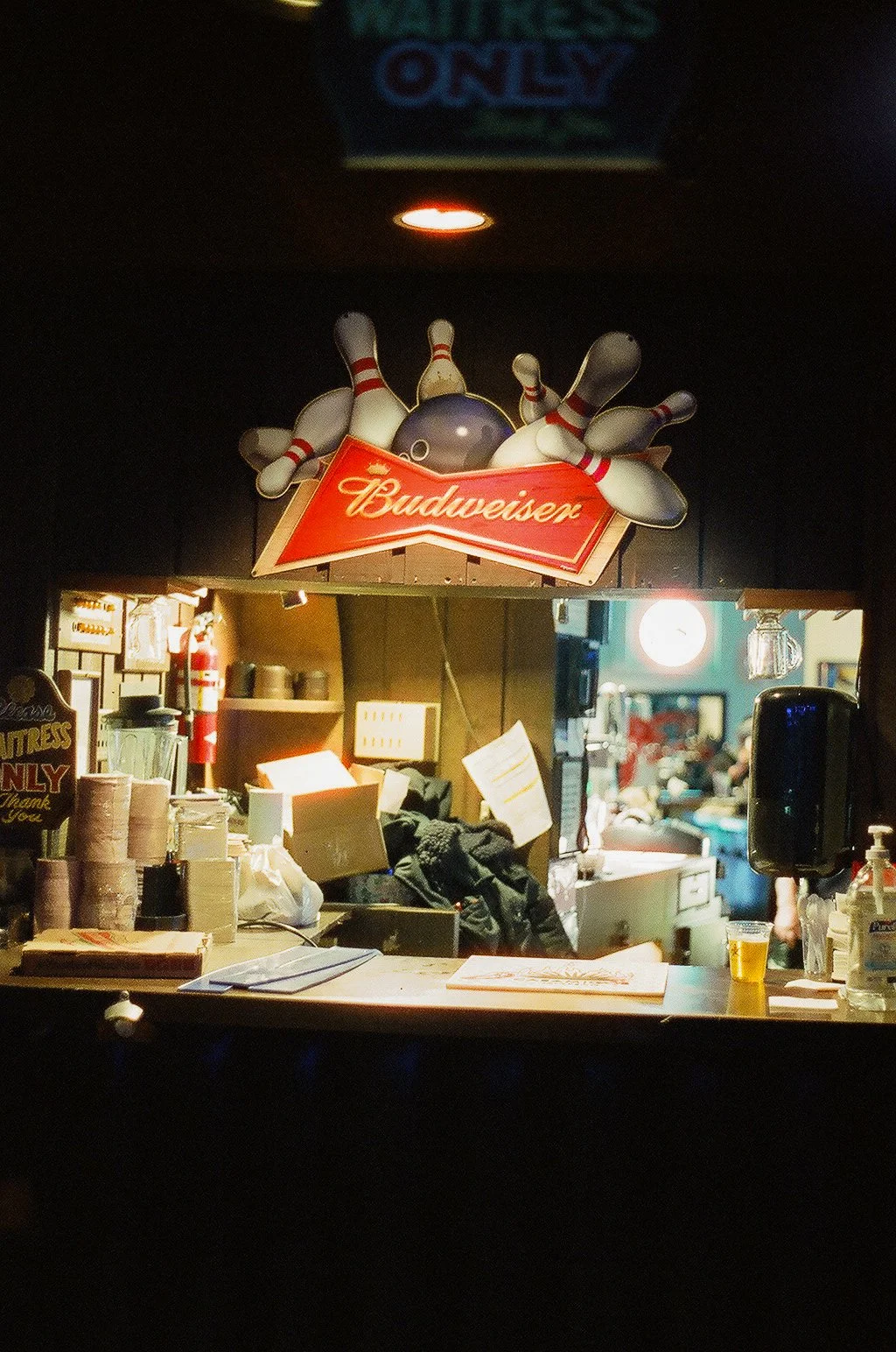 A view of a bar or counter area with a neon sign above that says 'Budweiser' with bowling pins and a bowling ball on top. Behind the counter are various supplies, a clock, and people in the background.