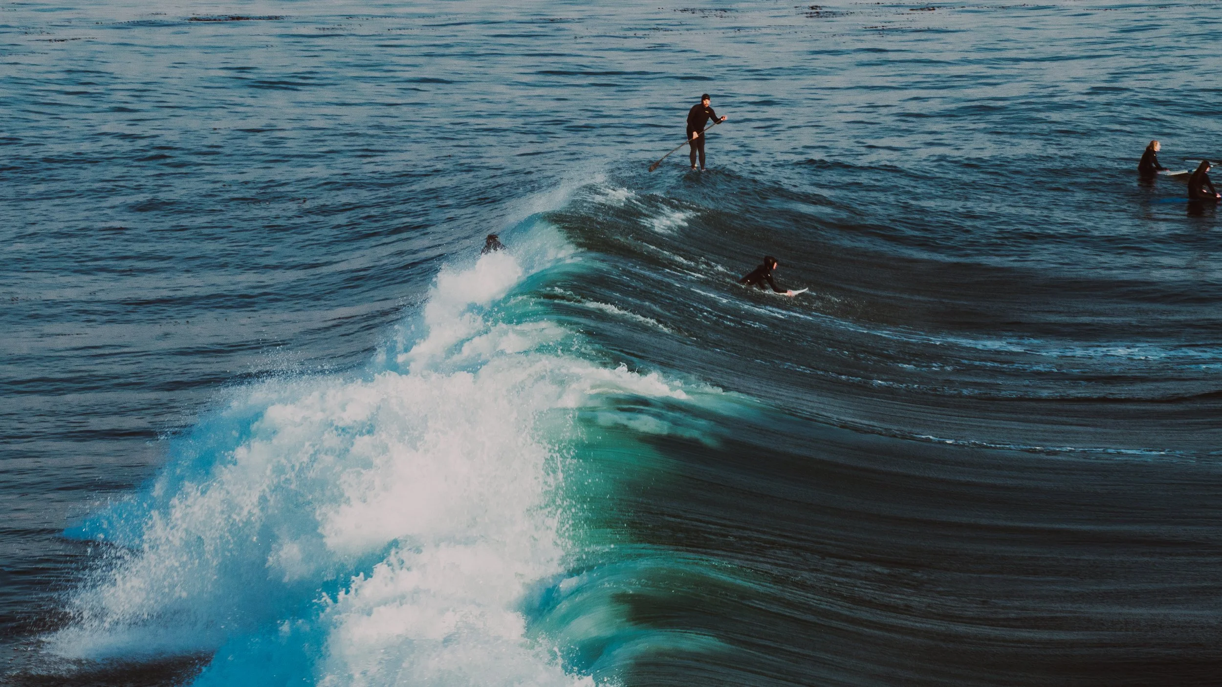People surfing on ocean waves, with one person standing on a paddleboard and several others riding surfing boards.