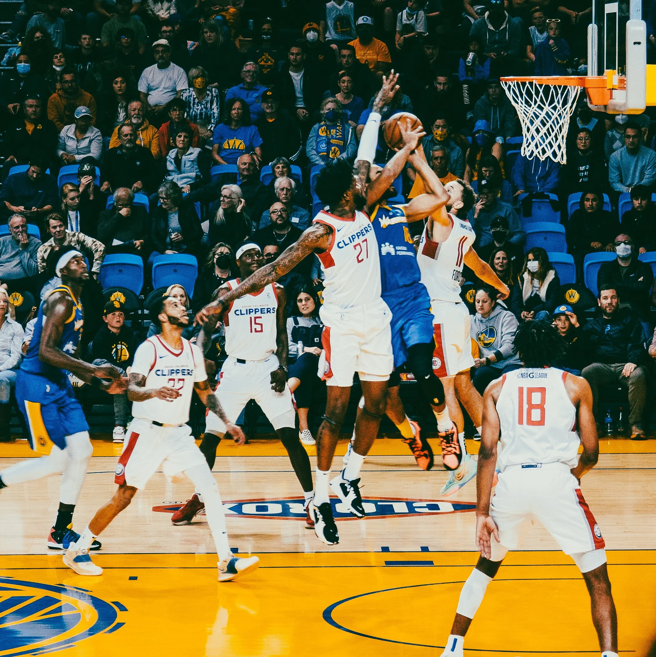 Basketball game with players jumping for the ball, surrounded by spectators in the stands.