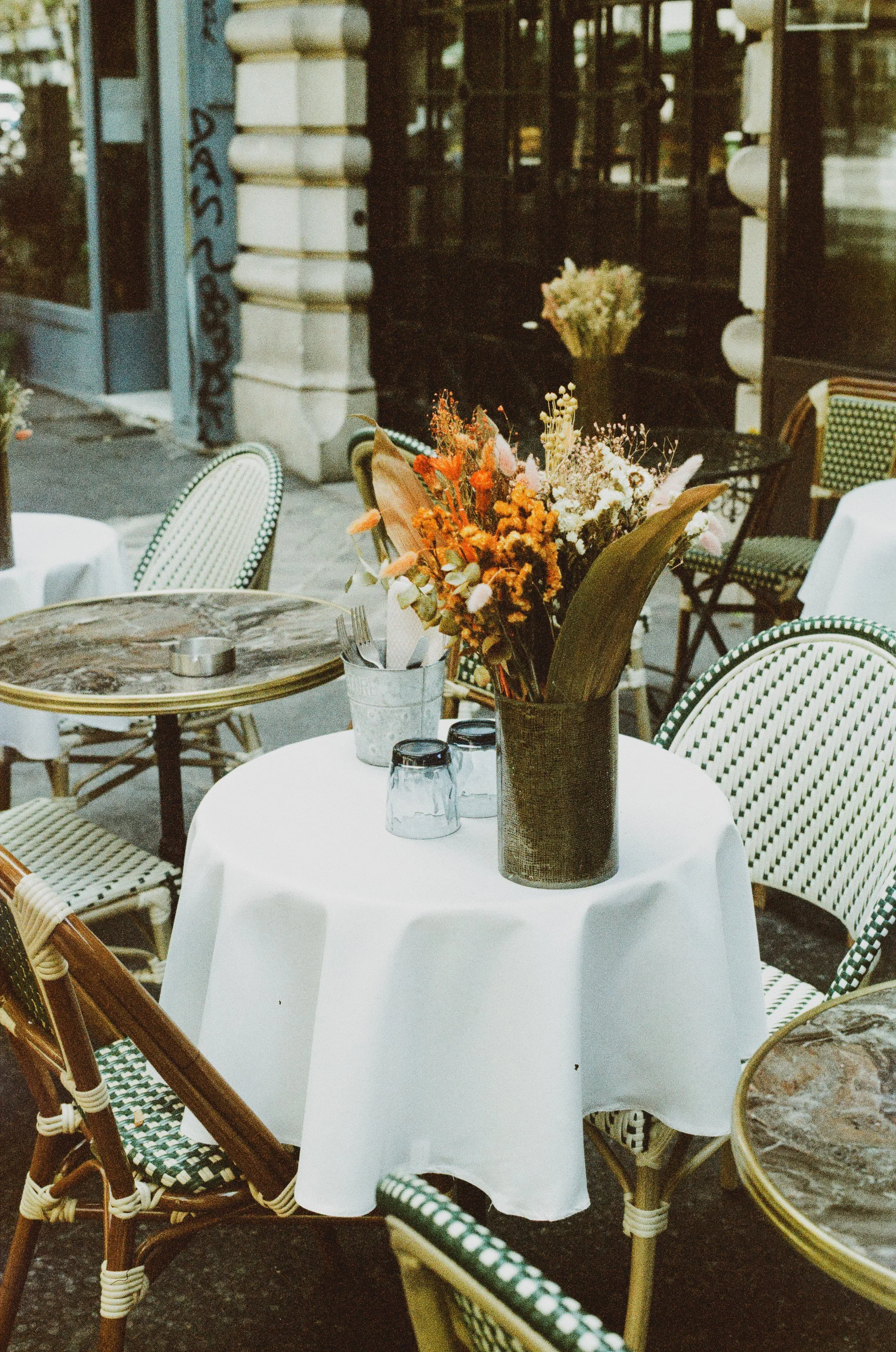 Outdoor cafe table with a white tablecloth, a large flower arrangement, and two glasses.