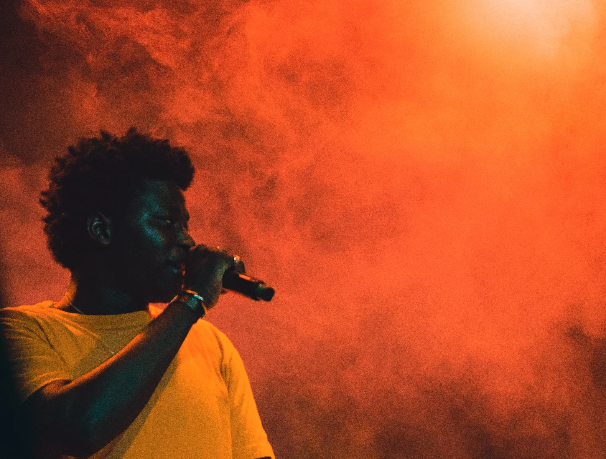 A person with dark skin and short curly hair singing into a microphone on stage, with orange and red lighting and smoke in the background.