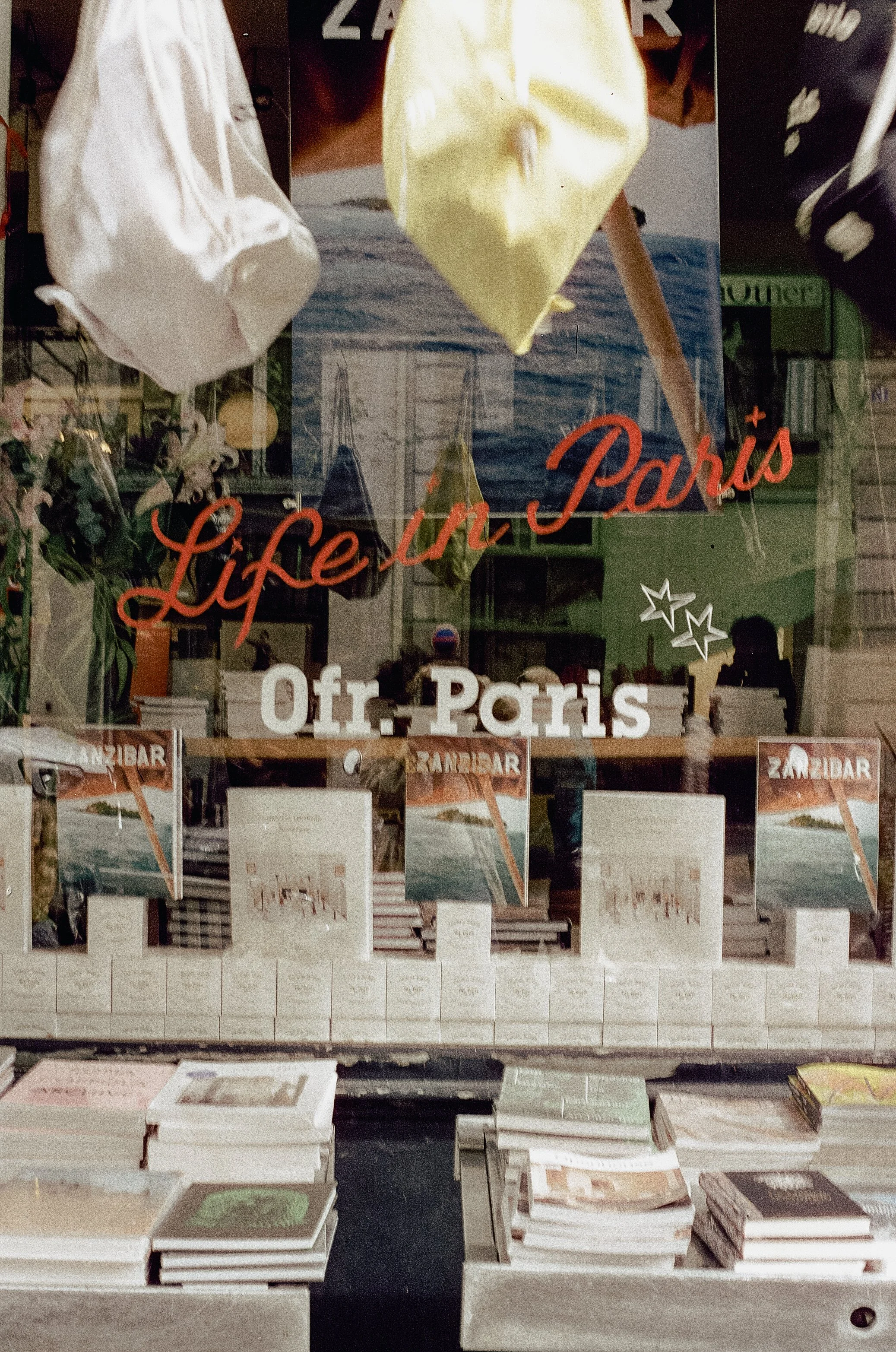 Books and posters displayed in a shop window with the words 'Life in Paris' and 'Off. Paris' written on the glass.