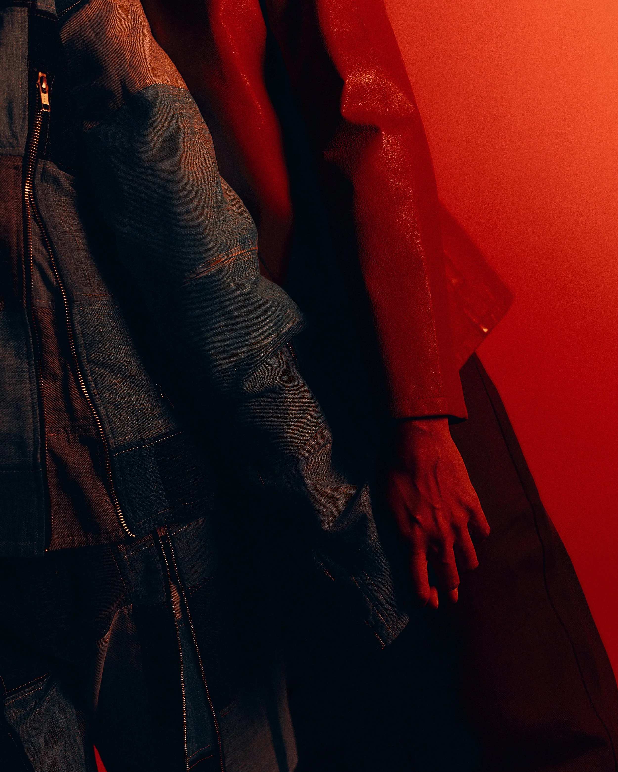 Close-up of a person's hand resting on their thigh, wearing dark clothing and a red leather jacket in a dimly lit setting with red lighting.