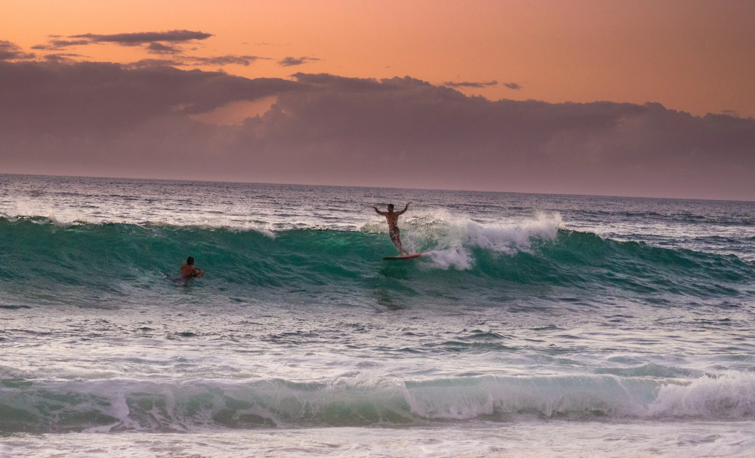 Man surfing a wave at sunset, another swimmer in the ocean.