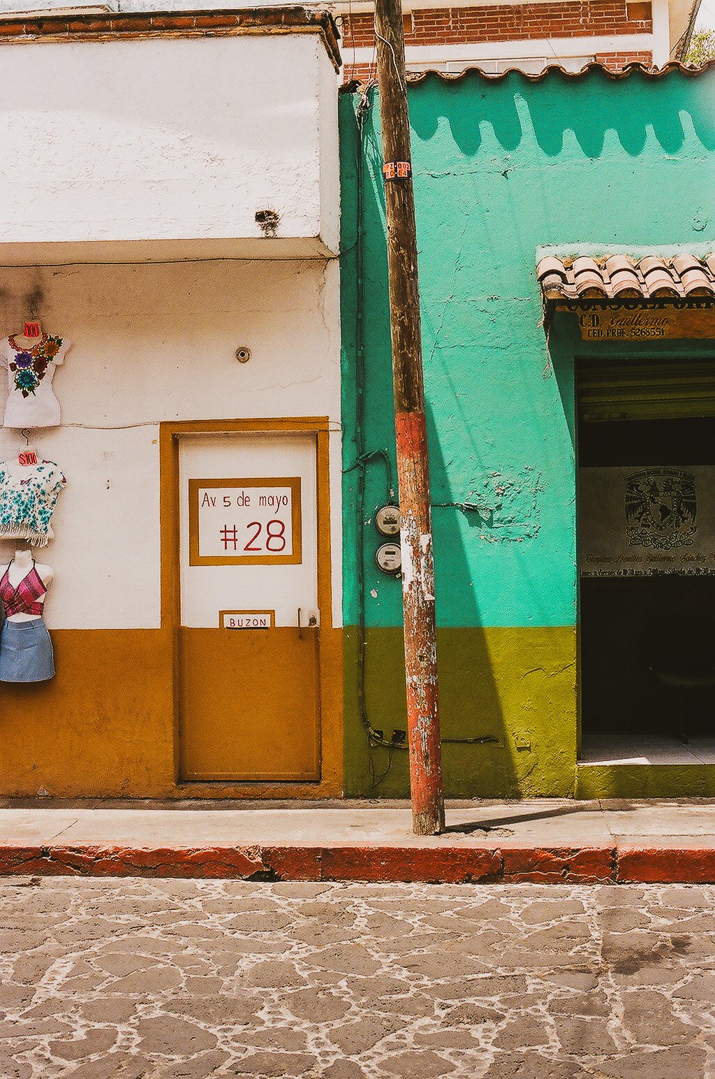 Colorful building with white, green, and brown walls, a wooden door with a sign reading 'Av. 5 de mayo #28', and a small sign below labeled 'BUZON'. A rusty pole is attached to the building near the door, and the street pavement in the foreground is 