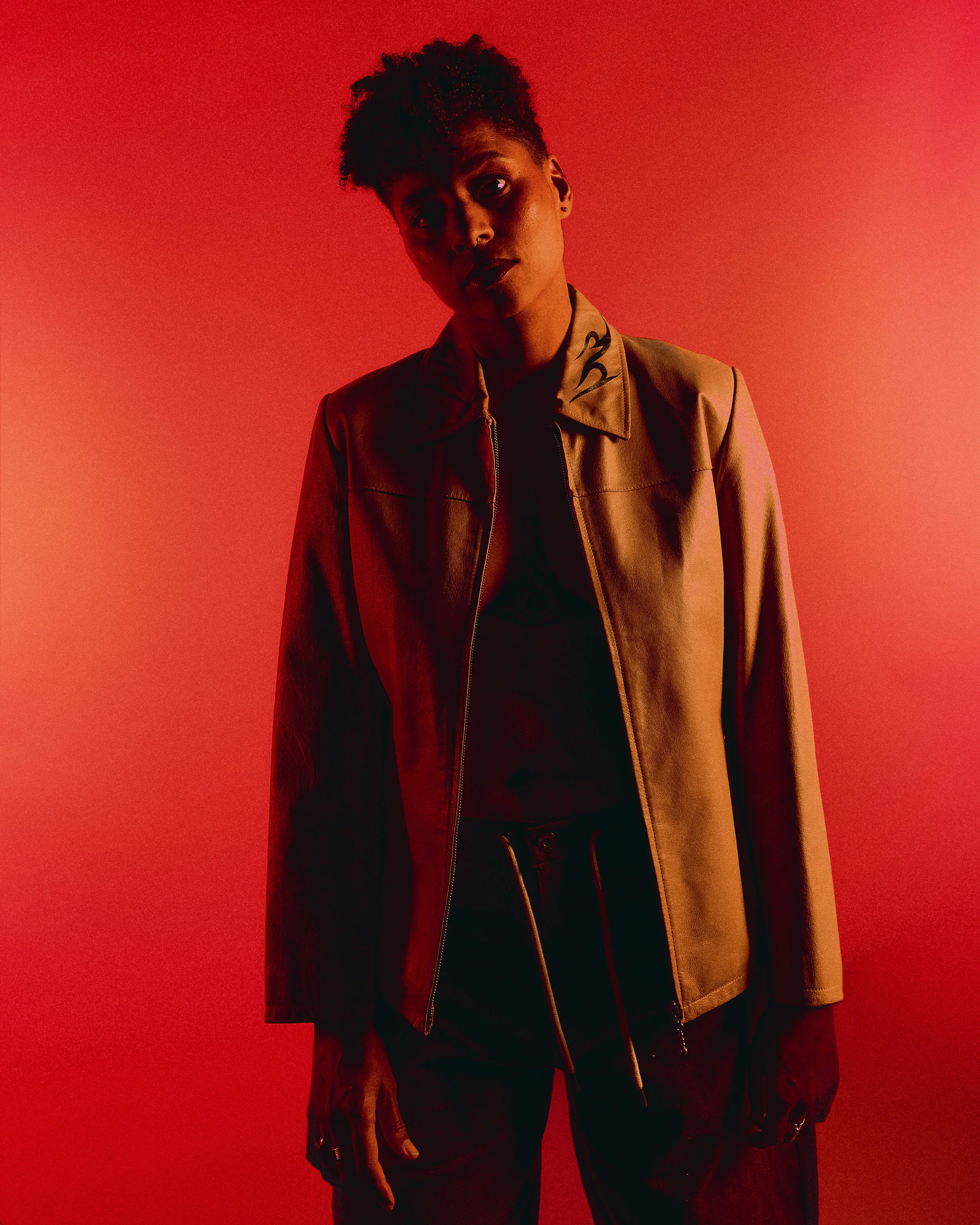 A woman in a brown leather jacket standing against a red background, with dramatic lighting casting shadows on her face.