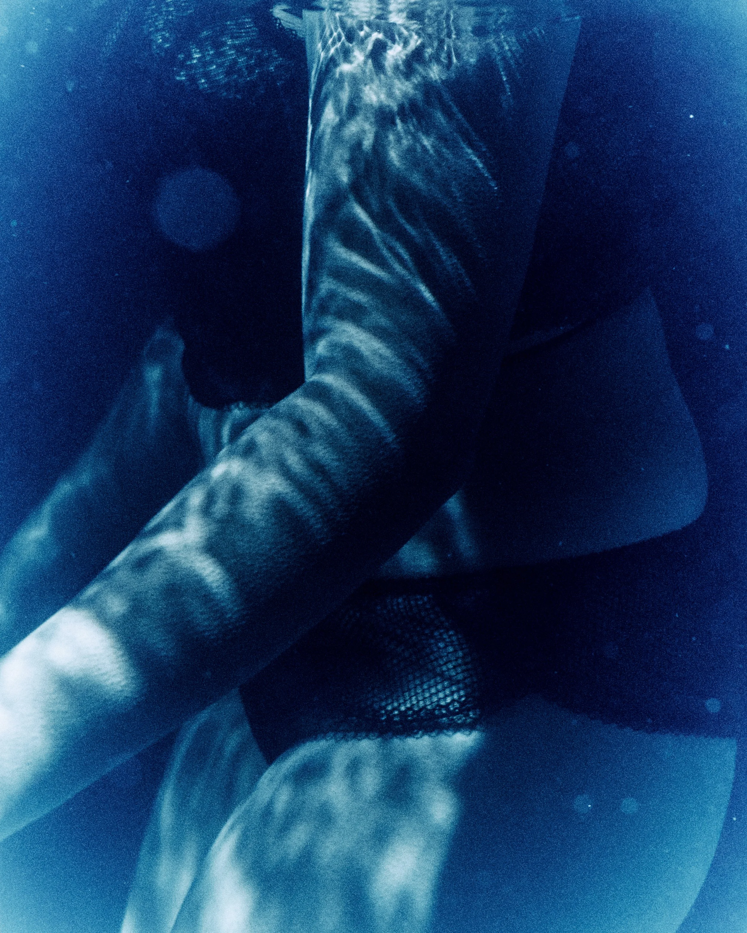 An underwater photograph of a person with short hair, wearing a black outfit, crouching with arms bent and close to the body.