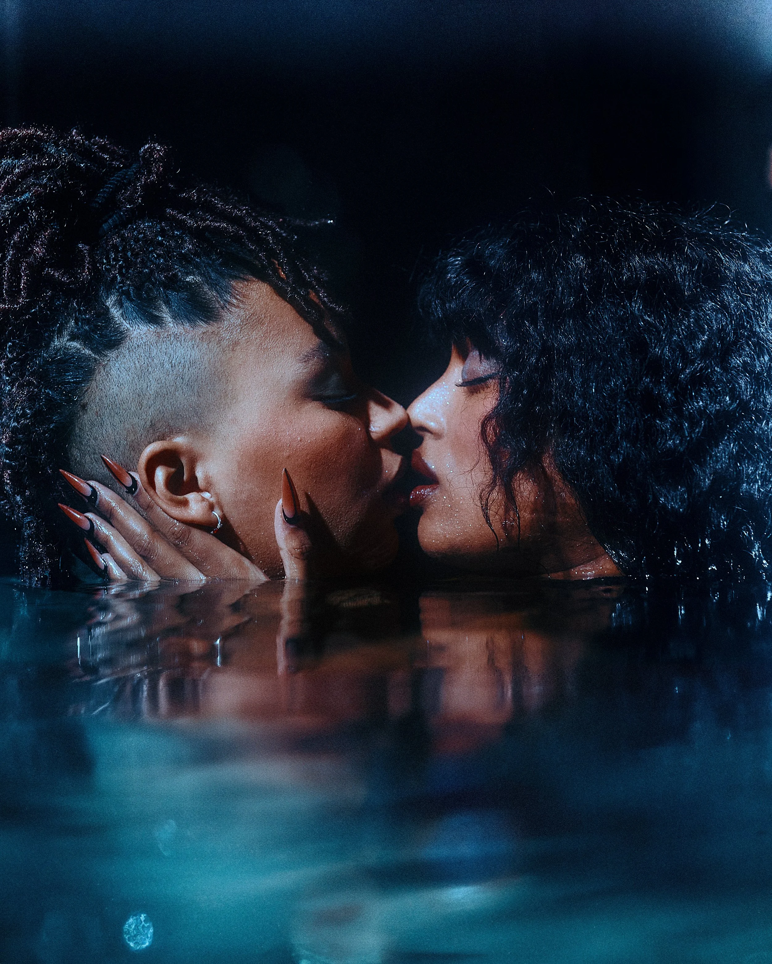 Two women with wet hair are about to kiss each other in a water setting, with dark background.