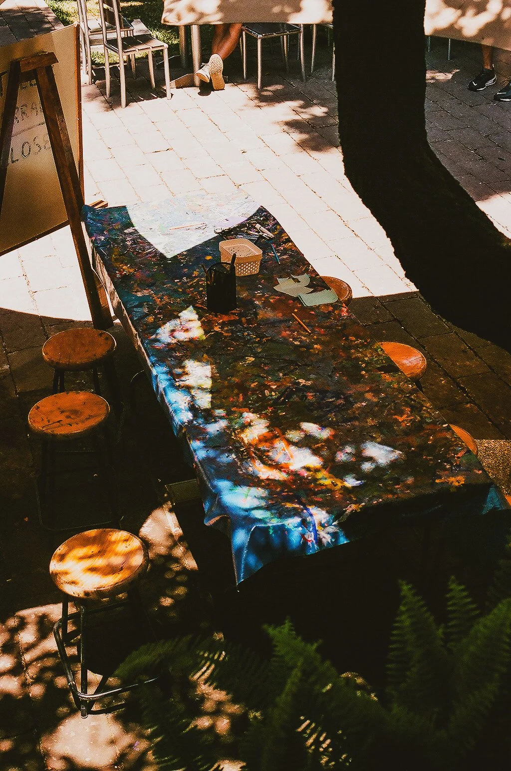 An outdoor art studio or workspace with a colorful, abstract painted tablecloth covering a long table. There are four wooden stools around the table and an easel with a whiteboard nearby. Shadows of trees and sunlight create dappled lighting on the s