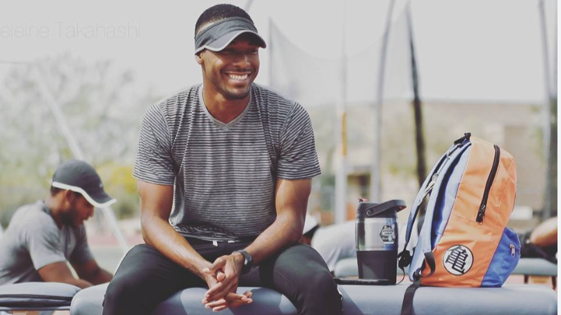 A man sitting outdoors smiling, wearing a cap and athletic clothing, with a backpack and water bottle nearby, and other people in the background.