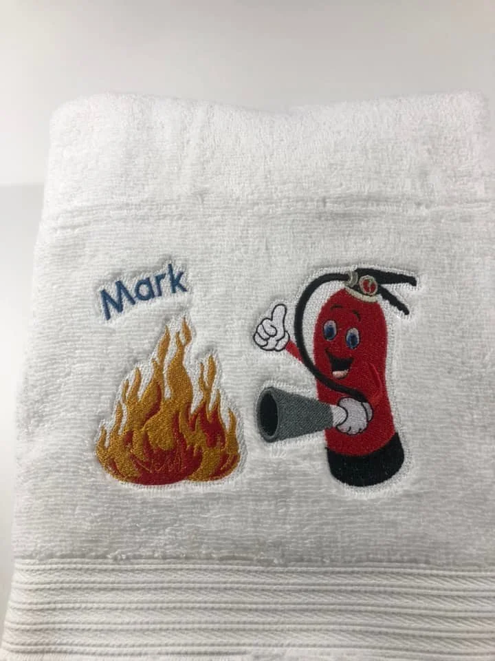 Embroidered white towel with a flaming fire patch and a cartoon red fire extinguisher character giving a thumbs up, with the name 'Mark' embroidered on it.