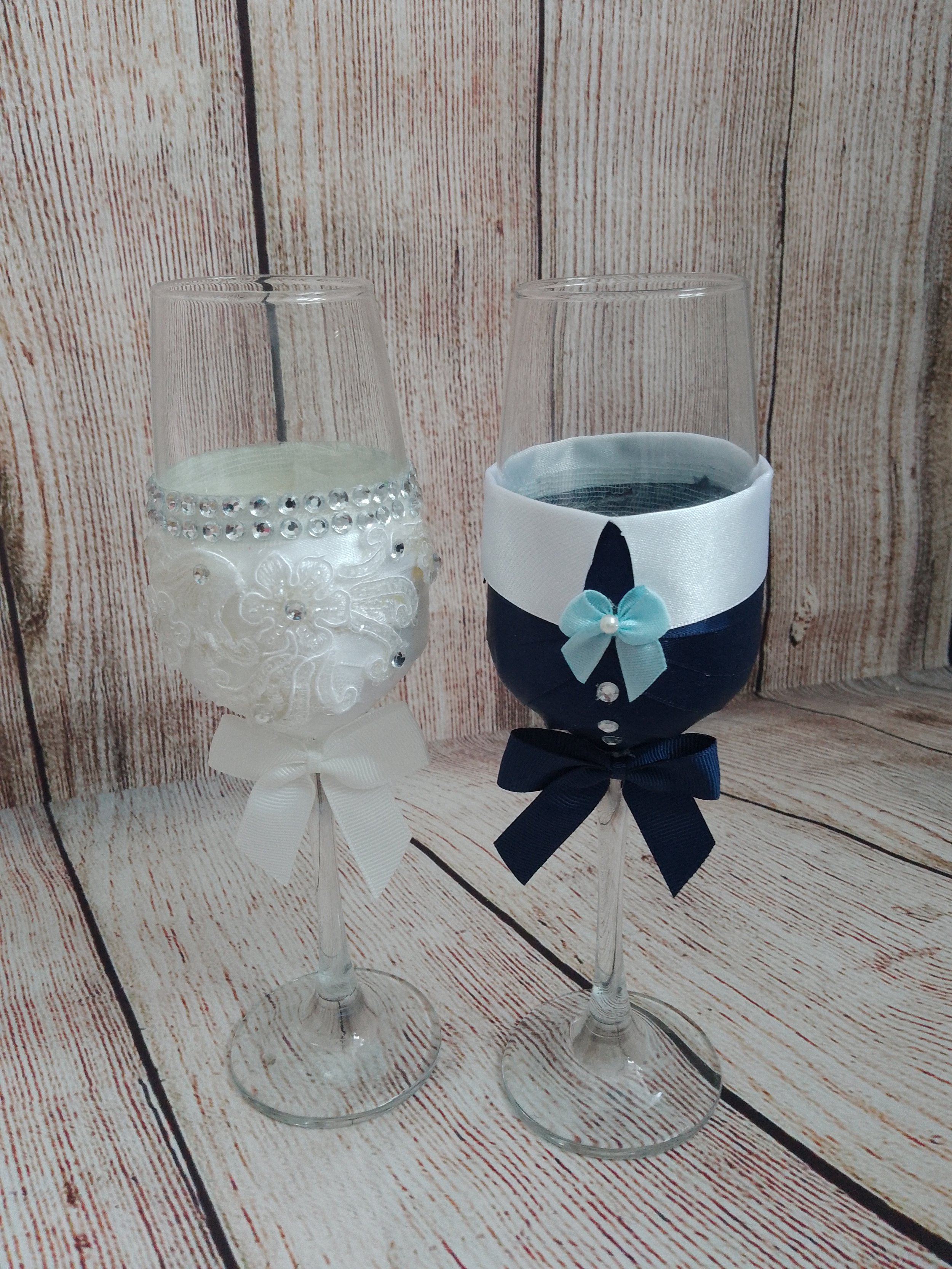Two champagne flutes decorated with fabric and ribbon, one with white embellishments and a white ribbon, the other with navy blue fabric, white ribbon, and a light blue bow with a pearl, set against a wooden background.