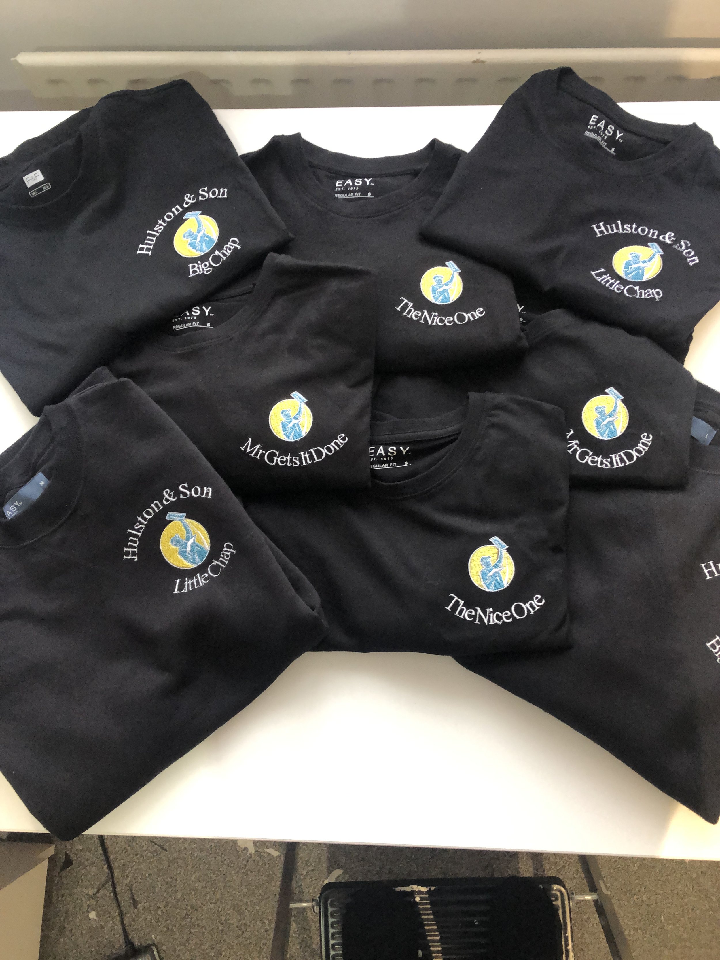 Multiple black T-shirts with a logo of a person in a blue shirt and yellow circle, and phrases like "Huston & Son Little Champ," "The Nice One," and "Mr Gets It Done" printed on them.