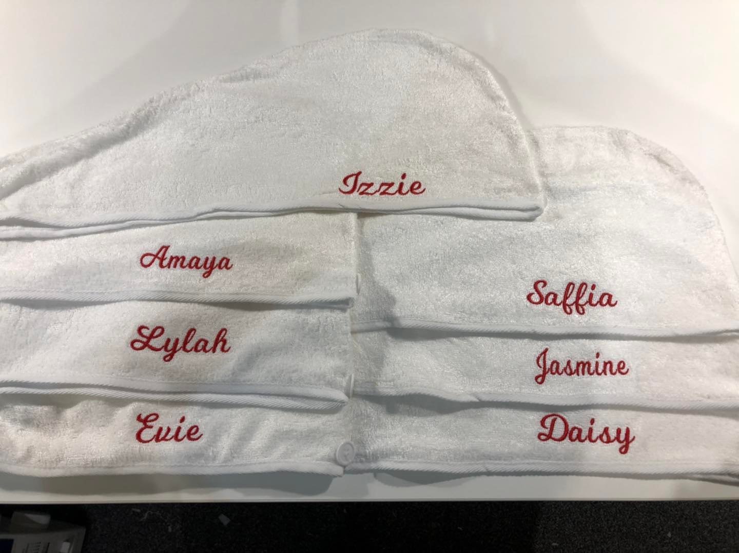 White towels with embroidered red names, including Izzie, Amaya, Saffia, Lylah, Jasmine, Evie, and Daisy, folded and displayed together.