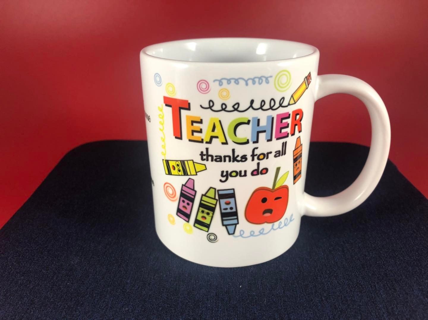 A white coffee mug with colorful text and illustrations. The text says "Teacher thanks for all you do" with various crayon and pencil drawings, including a red apple with a face, on a dark surface against a red background.