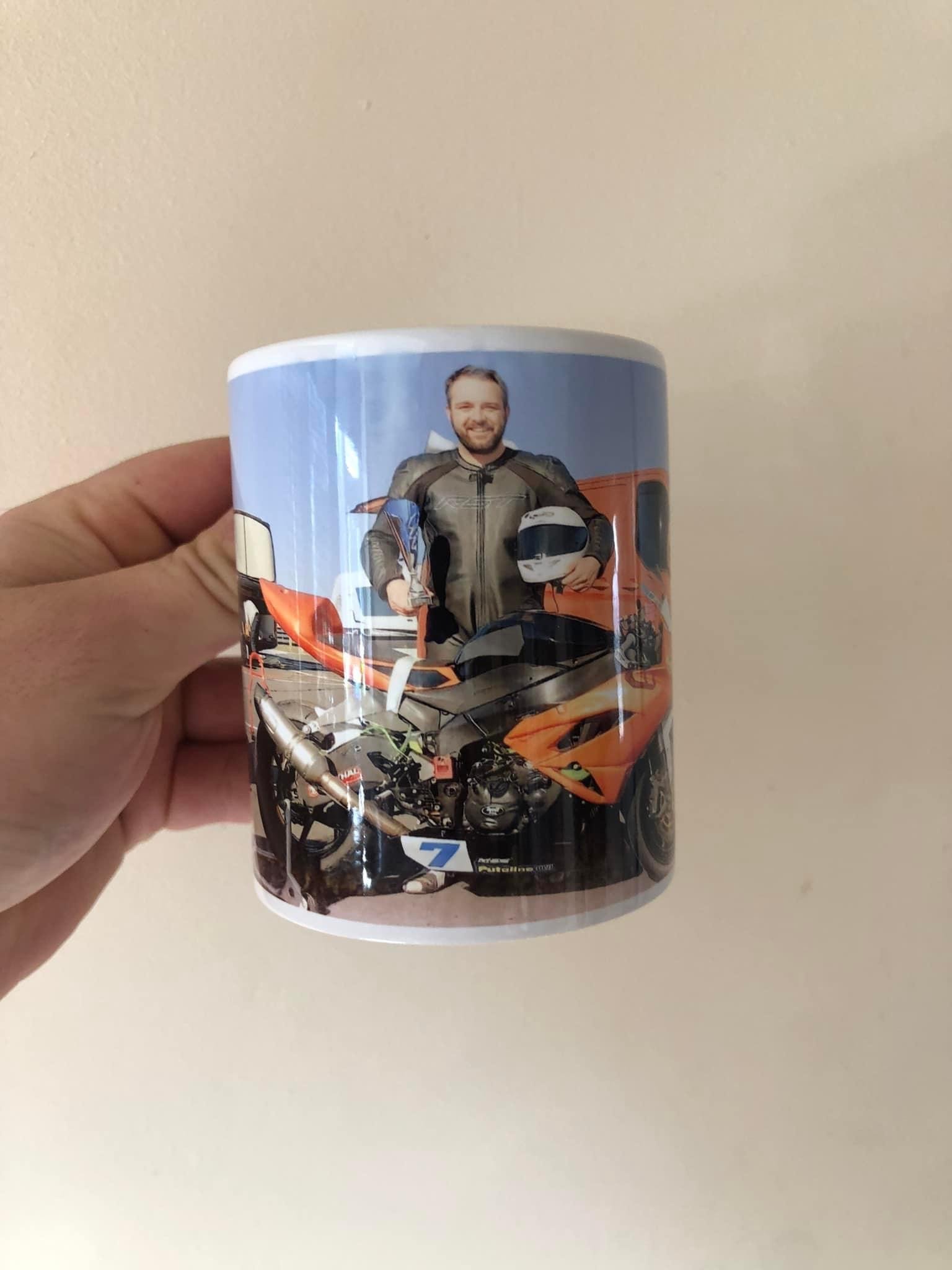 Person holding a mug with a photo of himself in a motorcycle gear, standing next to a motorcycle with an orange sports car in the background.