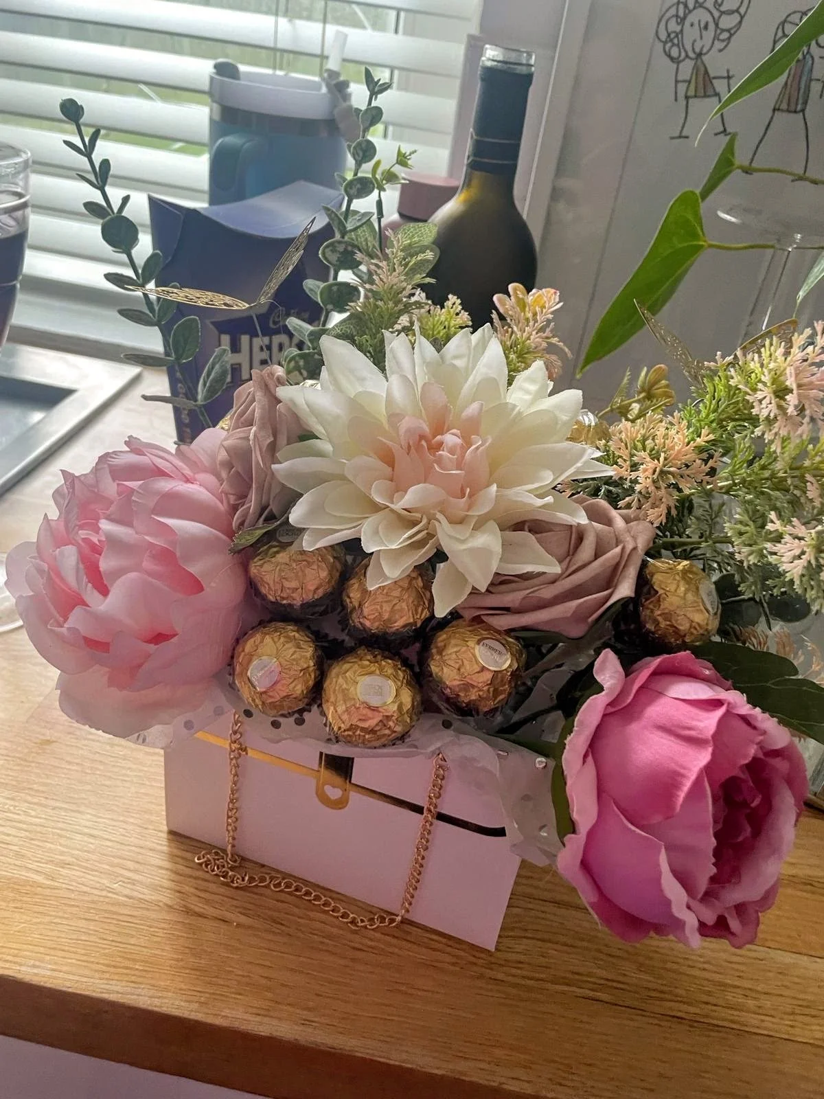 A flower arrangement with pink and white flowers, including peonies and roses, placed in a light pink box with gold chain accents on a wooden surface.