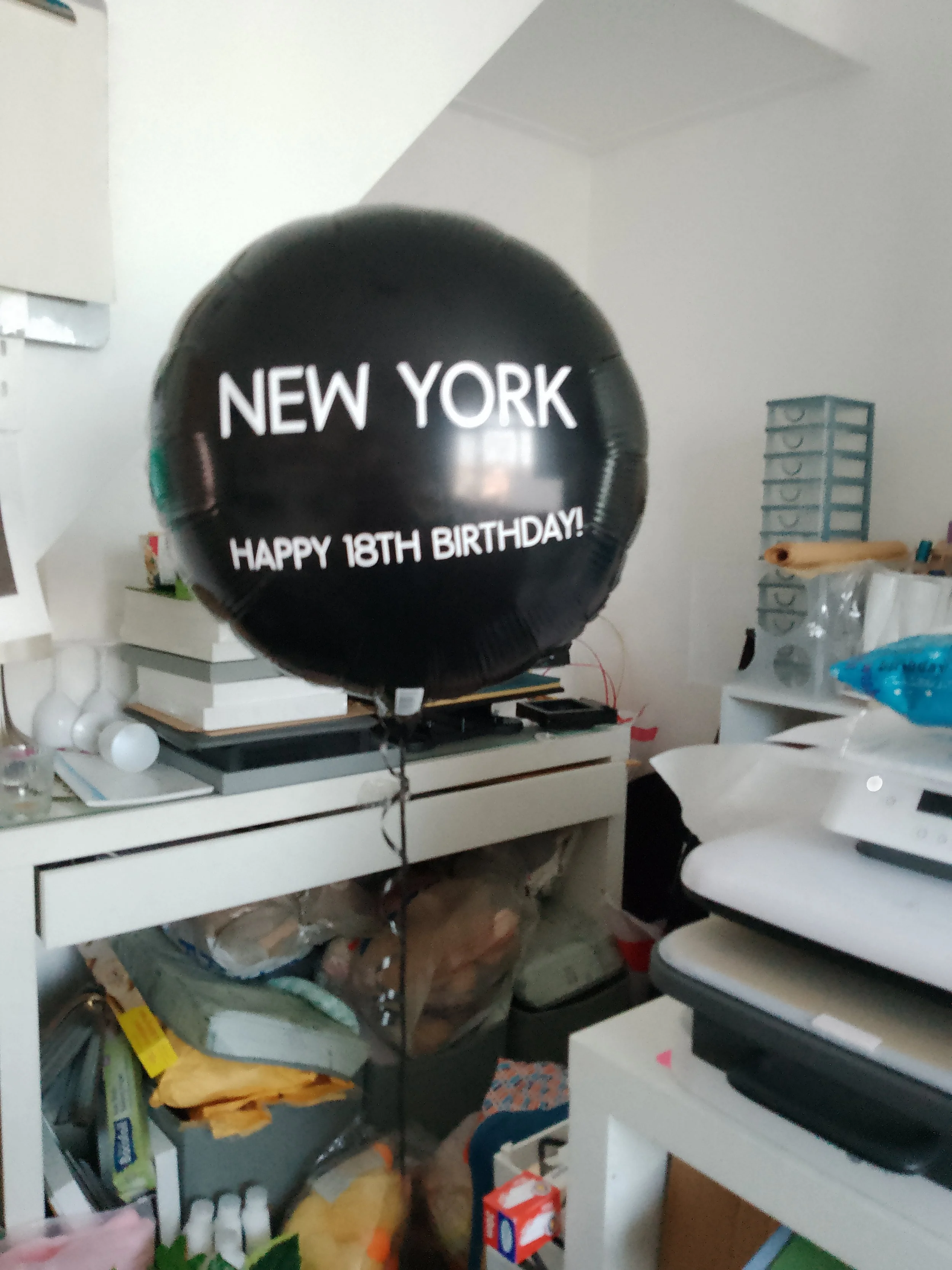 Black birthday balloon with white text reading "New York Happy 18th Birthday!" in a cluttered room.