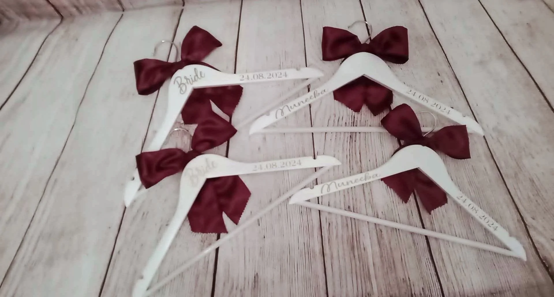 Set of four white wedding hangers with maroon bows, each engraved with different names and dates, placed on a light wooden floor.