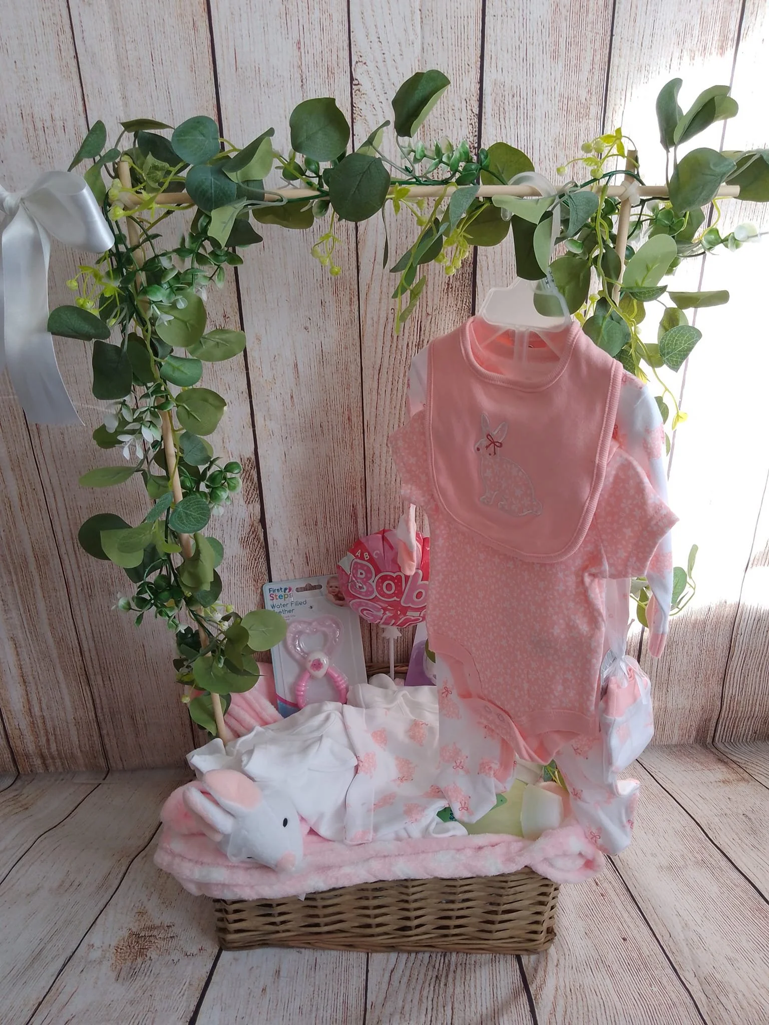 A baby gift basket with pink and white baby clothes, a bunny plush toy, pacifiers, and a 'Baby Girl' balloon, decorated with green foliage.