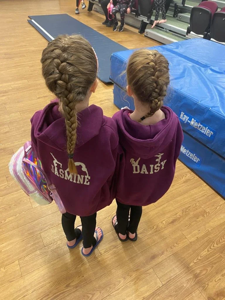 Two young girls with braided hair, wearing matching maroon hoodies with their names "Jasmine" and "Daisy" on the back, standing in an indoor gym. Jasmine's hoodie has a unicorn and horse design, and Daisy's has a unicorn design. Jasmine is carrying a