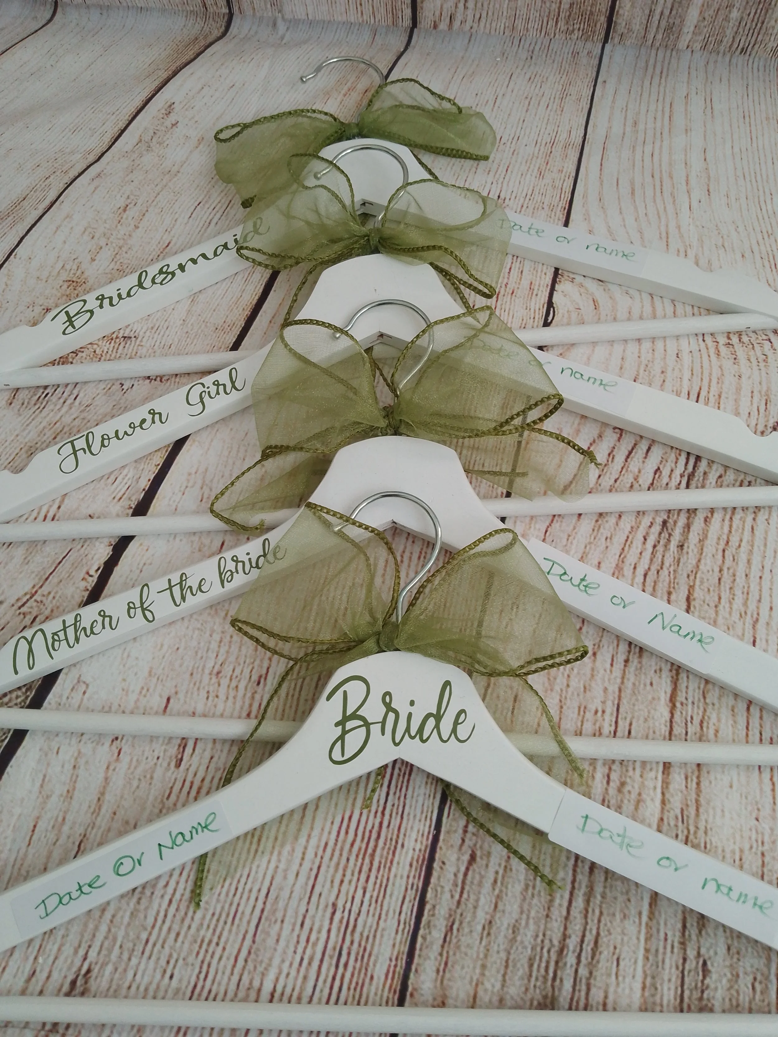 White wedding hangers with green bows and printed labels for bridal party roles, placed on a wooden surface. The hangers are labeled "Bride," "Mother of the bride," "Flower girl," and "Bridesmaid."