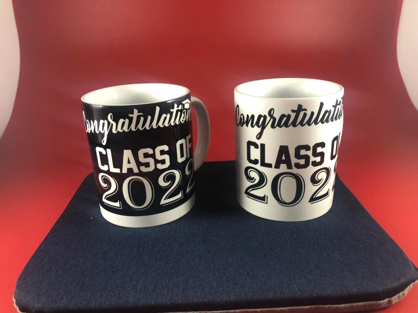 Two graduation mugs on a black surface with a red background; one black with white text, one white with black text, both saying 'Congratulations Class of 2022'.