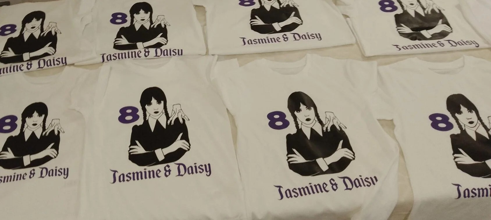 Multiple white t-shirts with a black and purple graphic of a woman with dark hair, crossed arms, and a confident expression. The shirts have text that reads "Jasmine & Daisy" and the number 8 in purple.