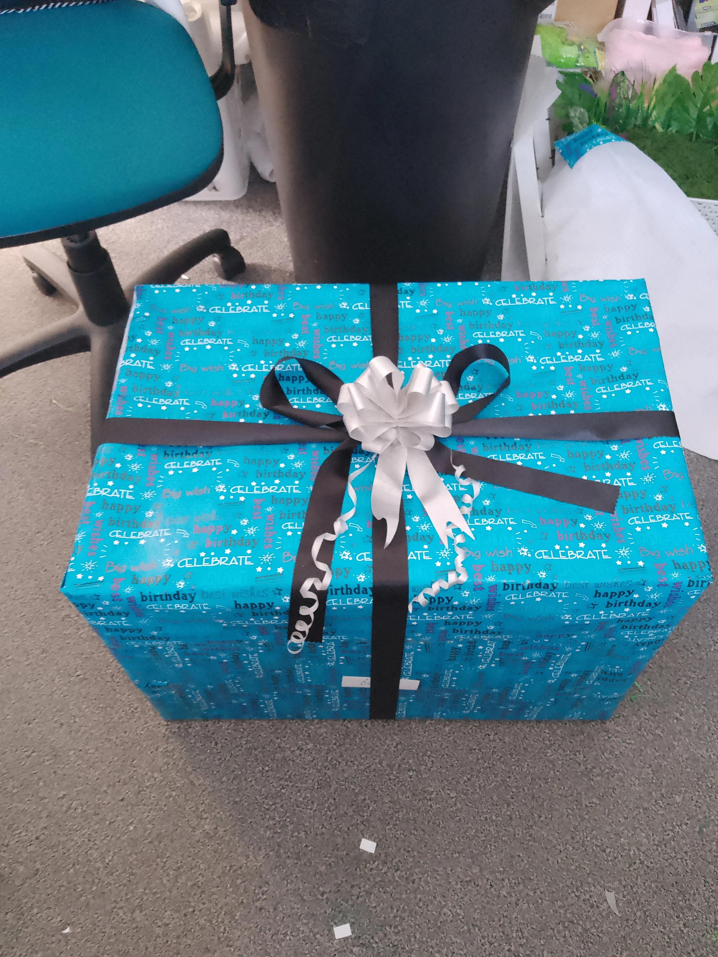 A decorated blue birthday gift box with a silver and black ribbon bow and curly ribbon on top, placed on a beige carpeted floor in an office or room setting, with a teal office chair, a black trash can, and miscellaneous items in the background.