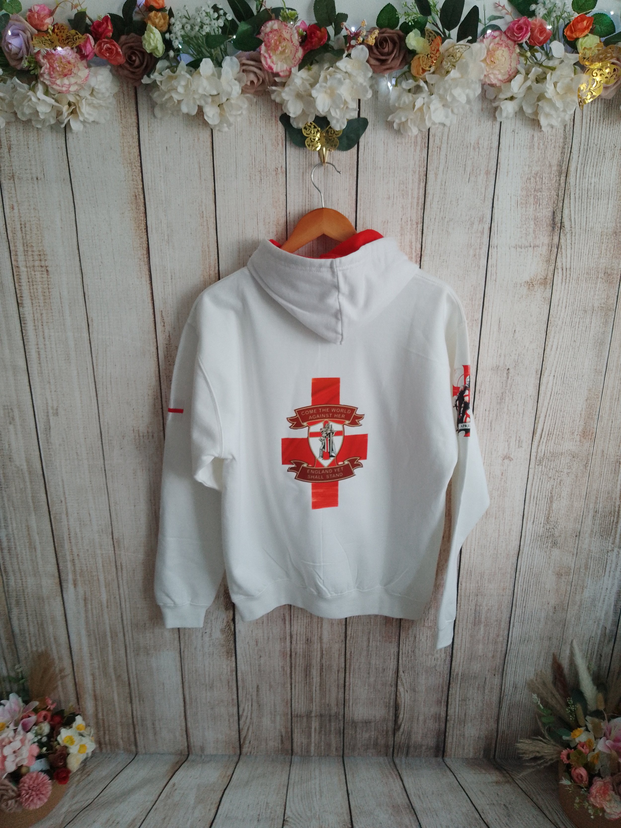 White hoodie with red cross and insignia on back hanging on a wooden hanger against a light wood paneled wall, decorated with flowers.