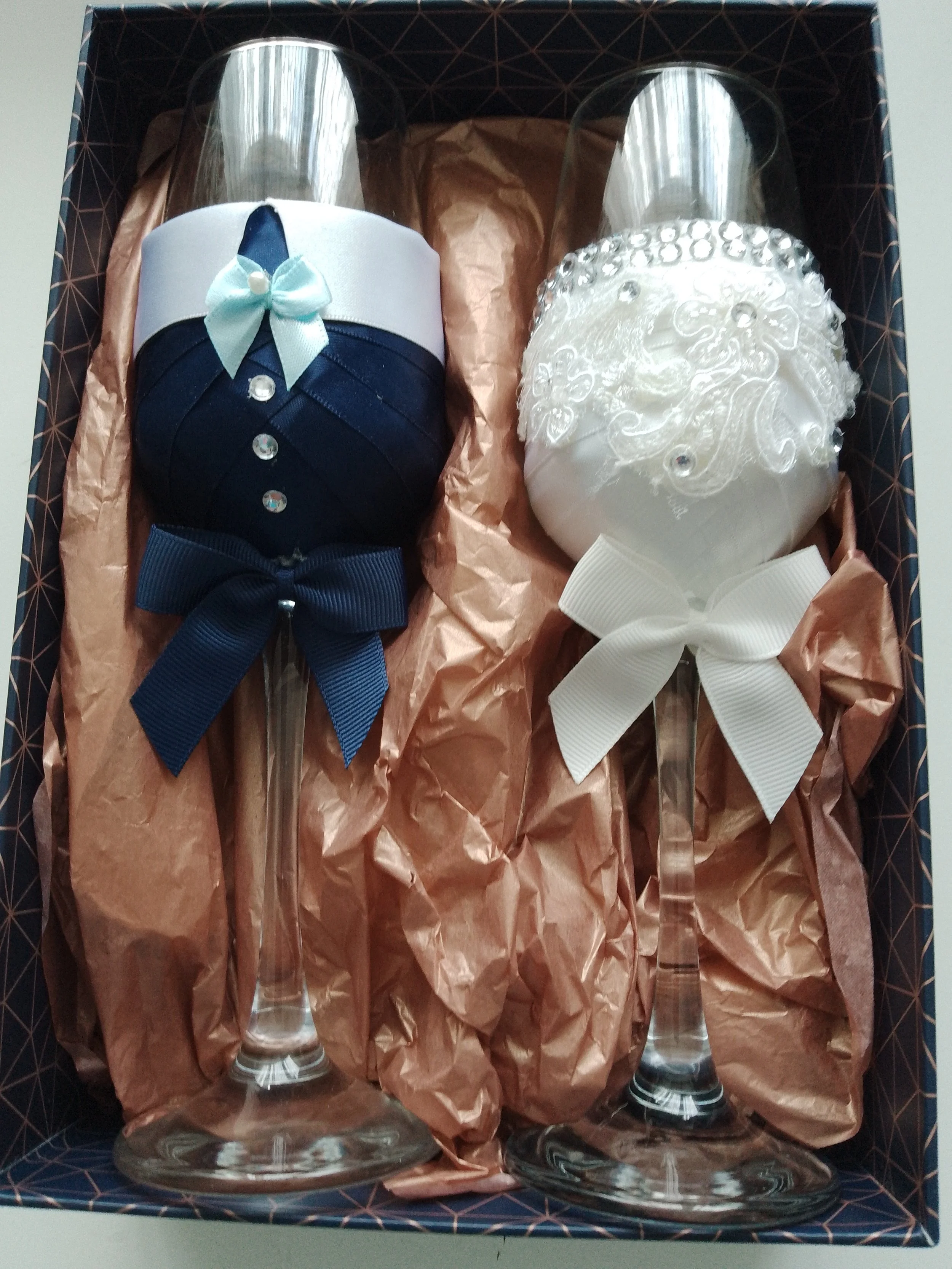A set of two decorative champagne flutes with fabric covers designed for a wedding or special occasion. The left flute is dressed as a groom, with a navy blue cover resembling a tuxedo, complete with a white collar, blue bow tie, and decorative butto