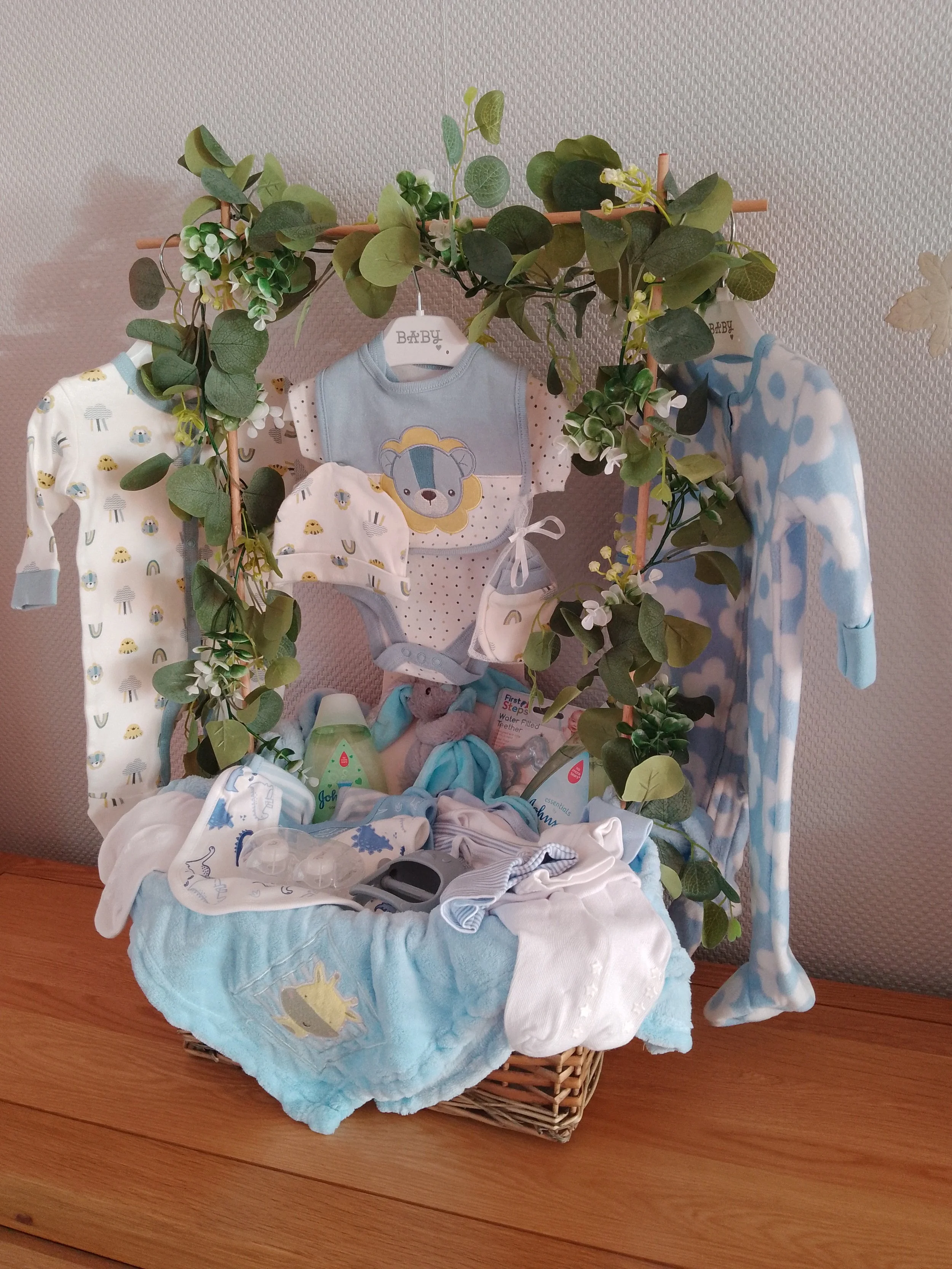 A baby ensemble display with hanging clothes, plush toys, bottles, and accessories in a wicker basket and a decorative leafy green garland on top.