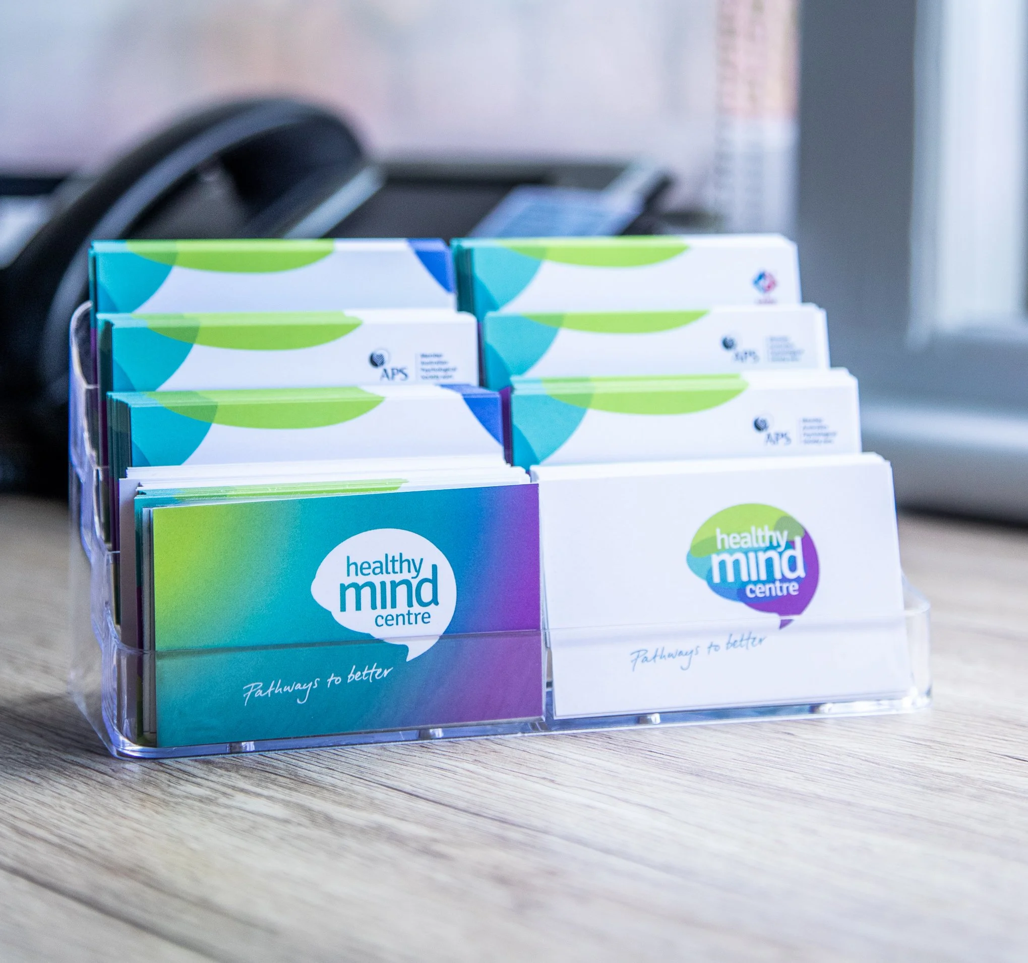 Business cards for Healthy Mind Centre in a clear plastic holder on a wooden desk.