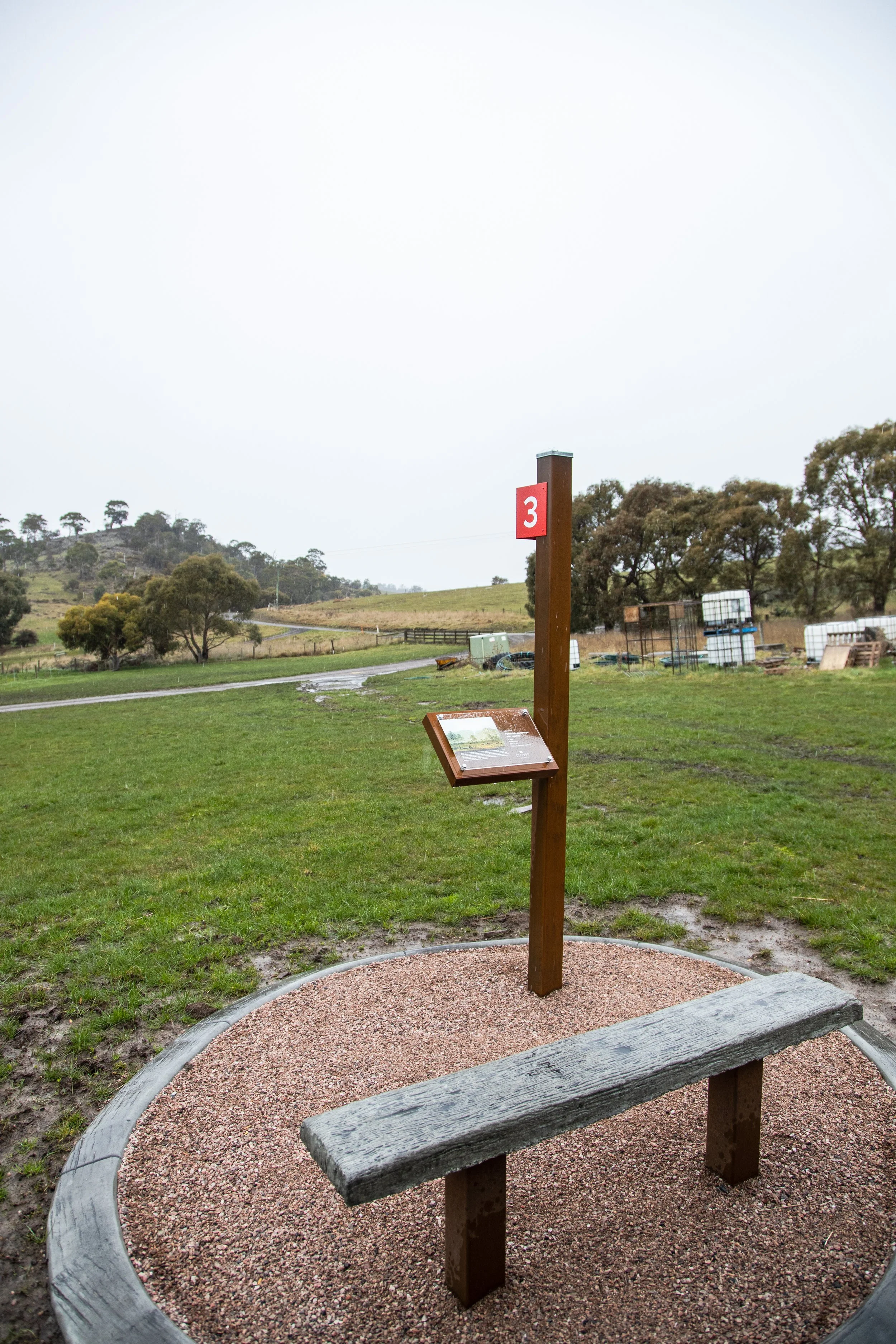 A wooden bench with a backrest sits in a shallow circular bed filled with small gravel. A post stands nearby with a red sign displaying the number 3 and an informational sign attached to it. The background is a rural landscape with grassy fields, tre