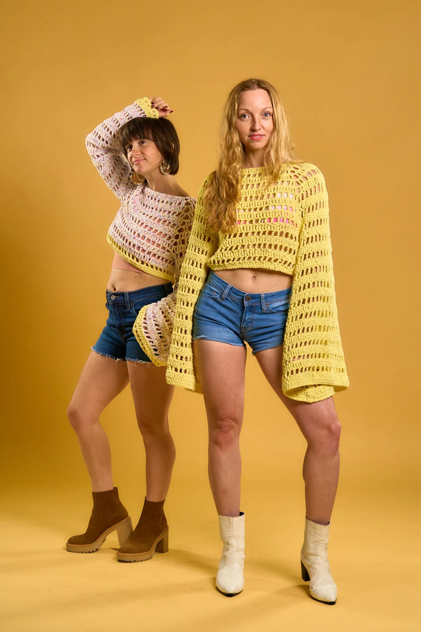 Two women with long hair wearing crochet tops and denim shorts, standing against a yellow background. The woman on the left has short dark hair and earrings, wearing brown boots and socks. The woman on the right has long blonde hair, white boots, and a yellow crochet top.
