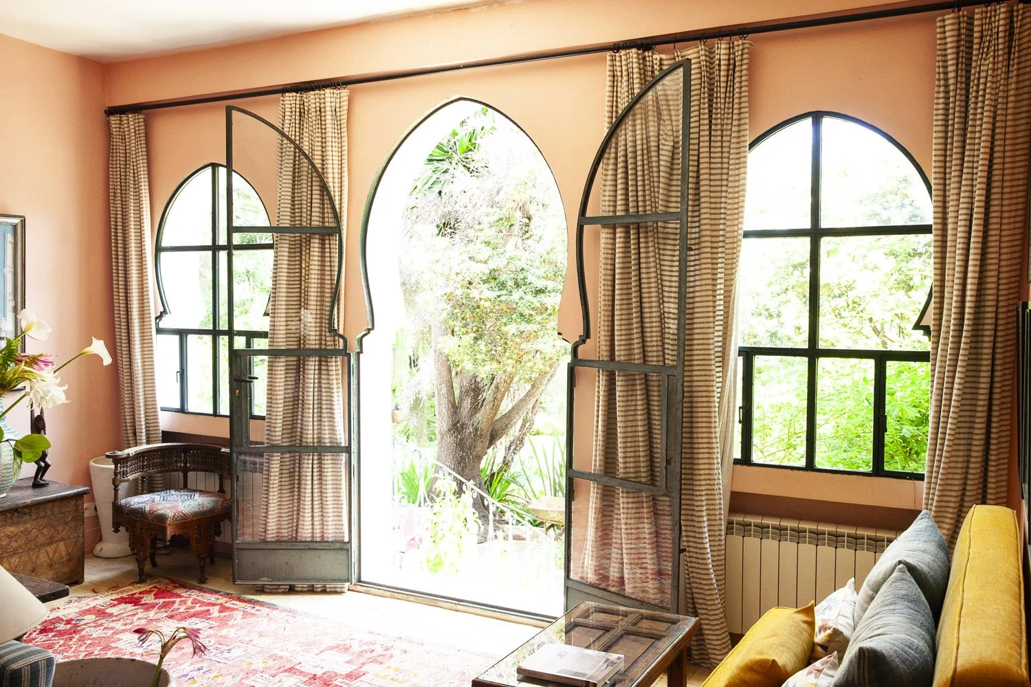 Living room with large arched windows and curtains, view of a garden with trees, wicker chair, floral arrangements, and a sofa with yellow and gray cushions.