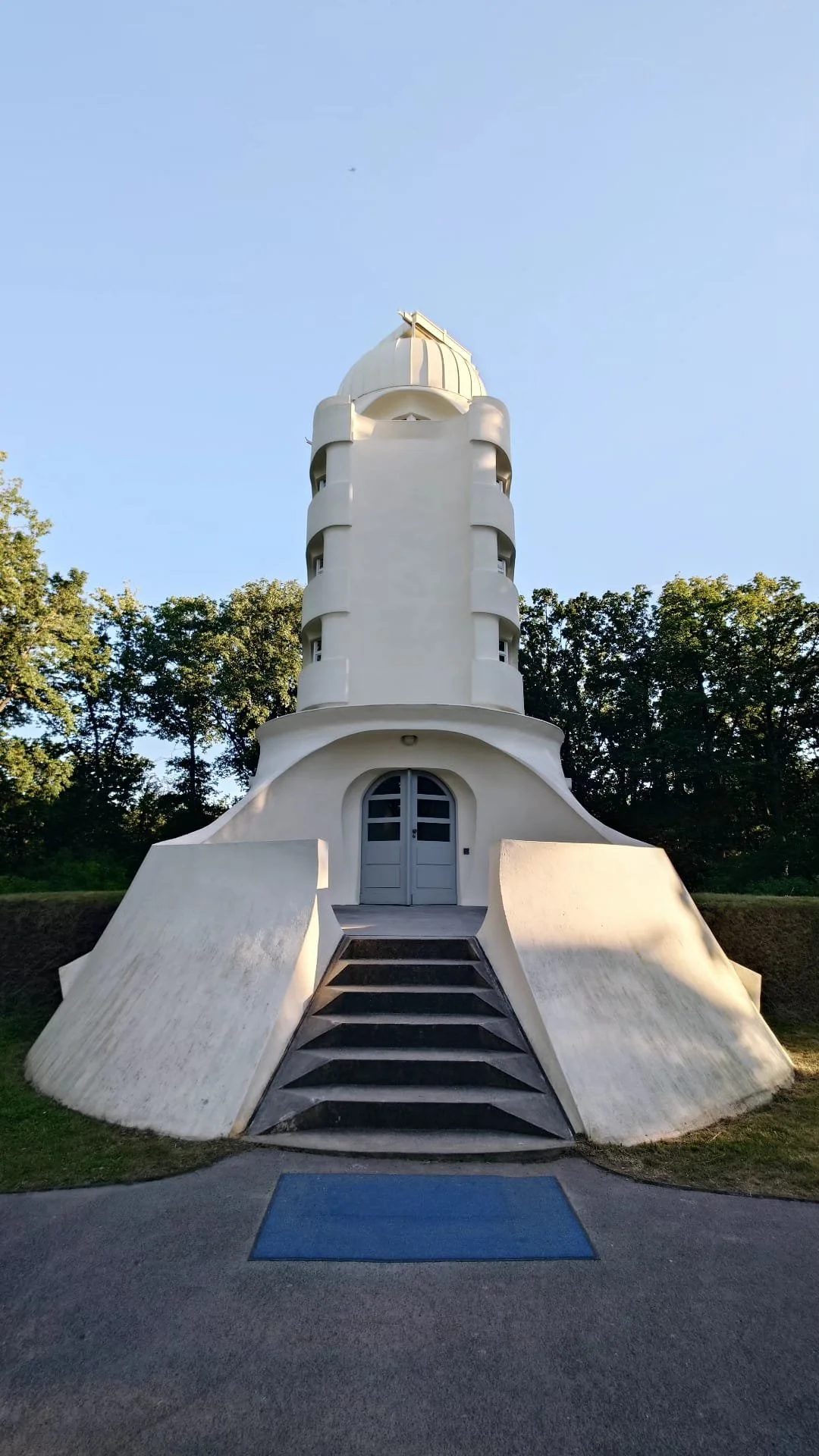 A tall, white, modern tower with rounded edges and multiple small windows, set against a backdrop of green trees and a blue sky.