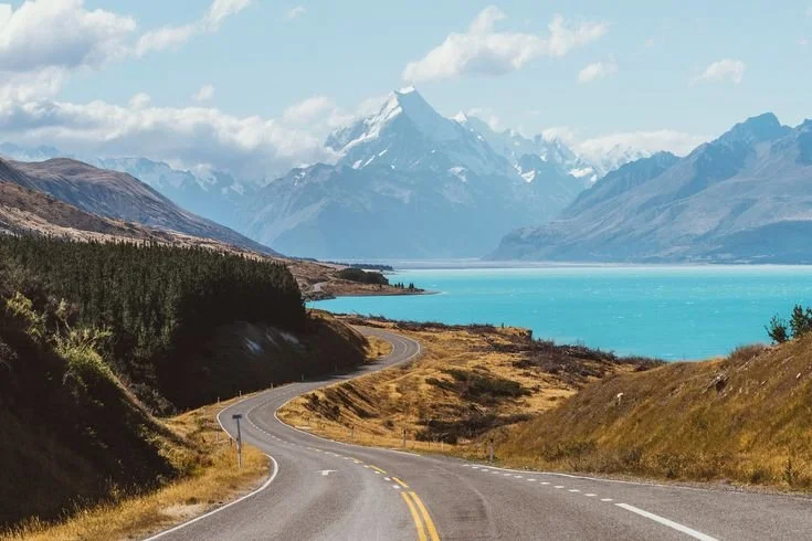 Winding road along a lake with mountains in the background.