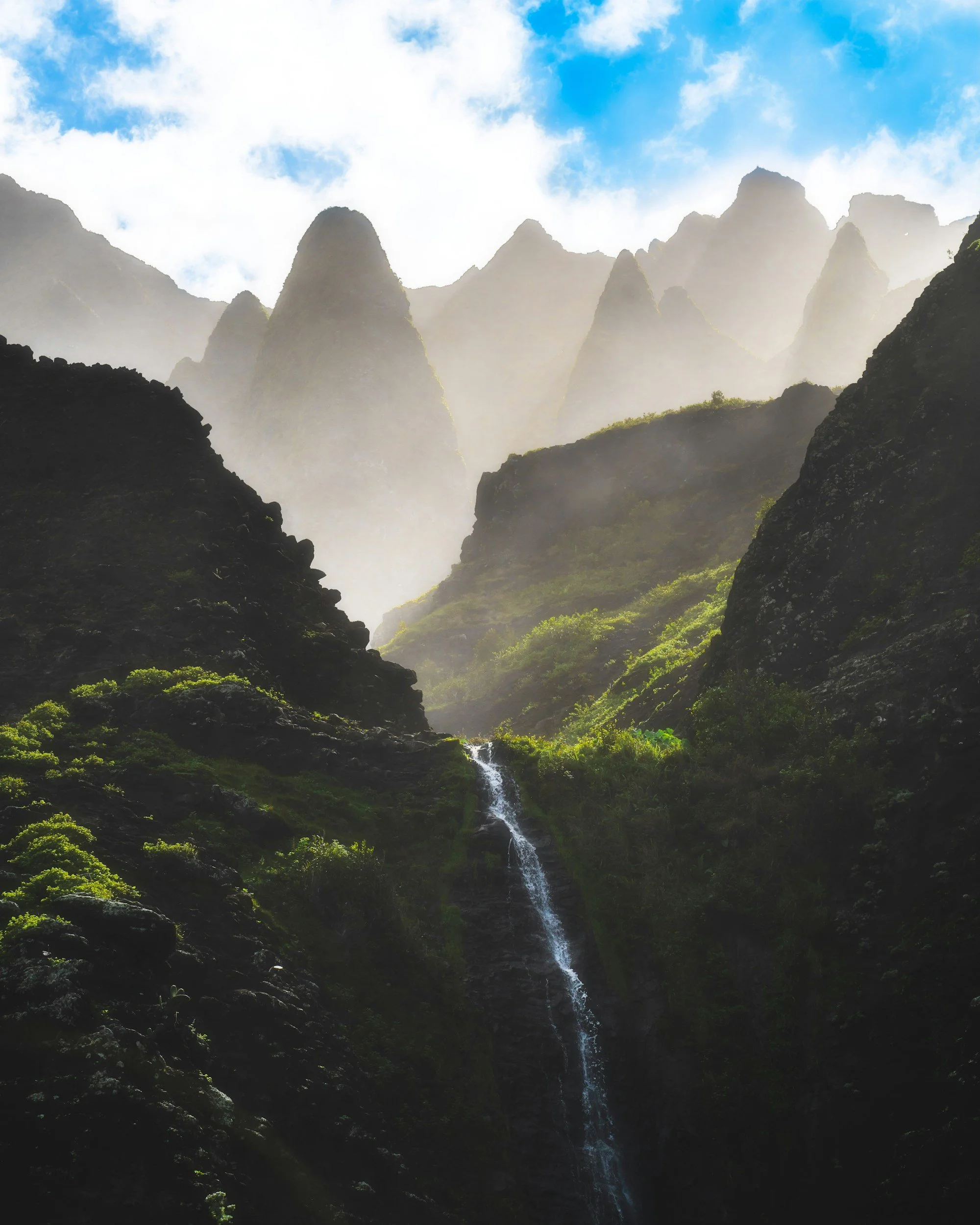 Mountains with mist and a waterfall in a lush green valley under a partly cloudy sky.