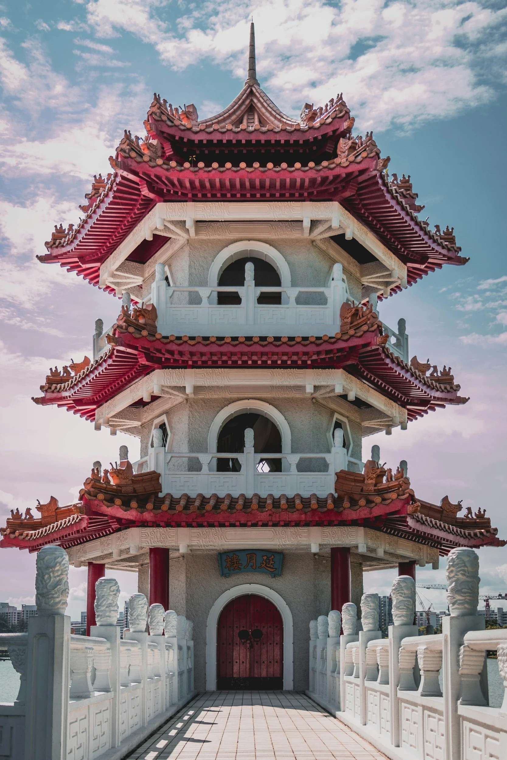 Traditional multi-tiered Chinese pagoda with red and white accents, ornate roof details, and decorative statues, set against a partly cloudy sky.