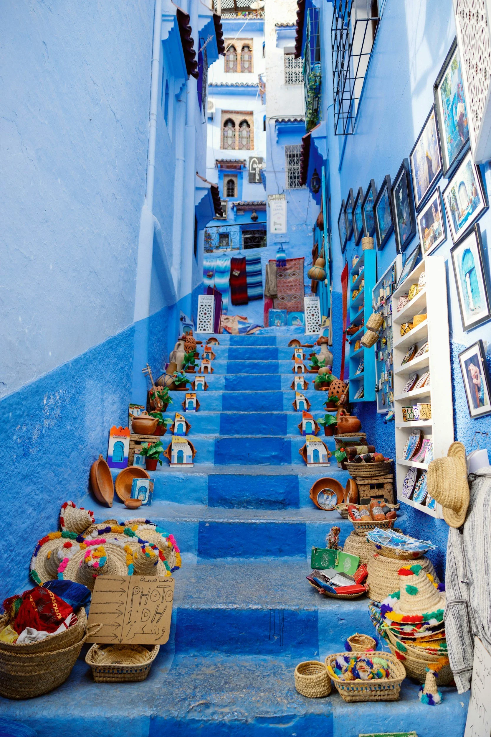 Blue-painted alleyway with stairs, decorated with pottery, woven hats, and artwork, in a Moroccan medina.