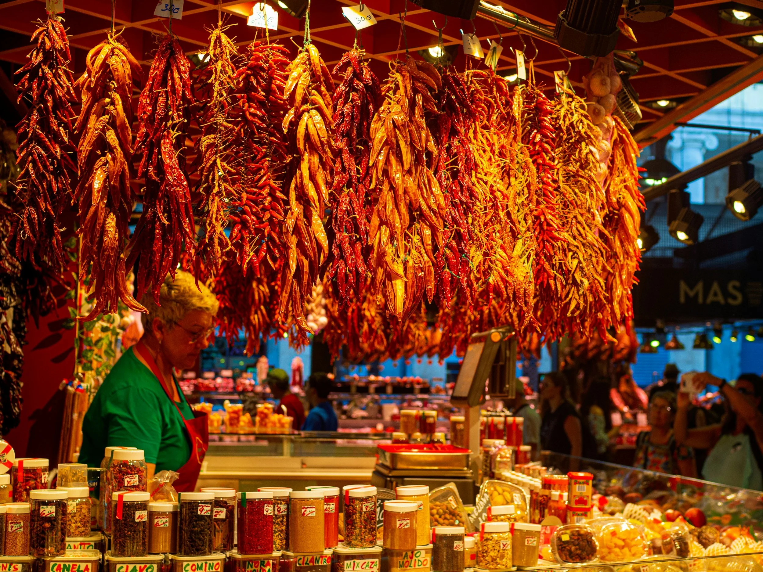 Market stall displaying hanging dried chili peppers and jars of various spices with shoppers and vendor in background.